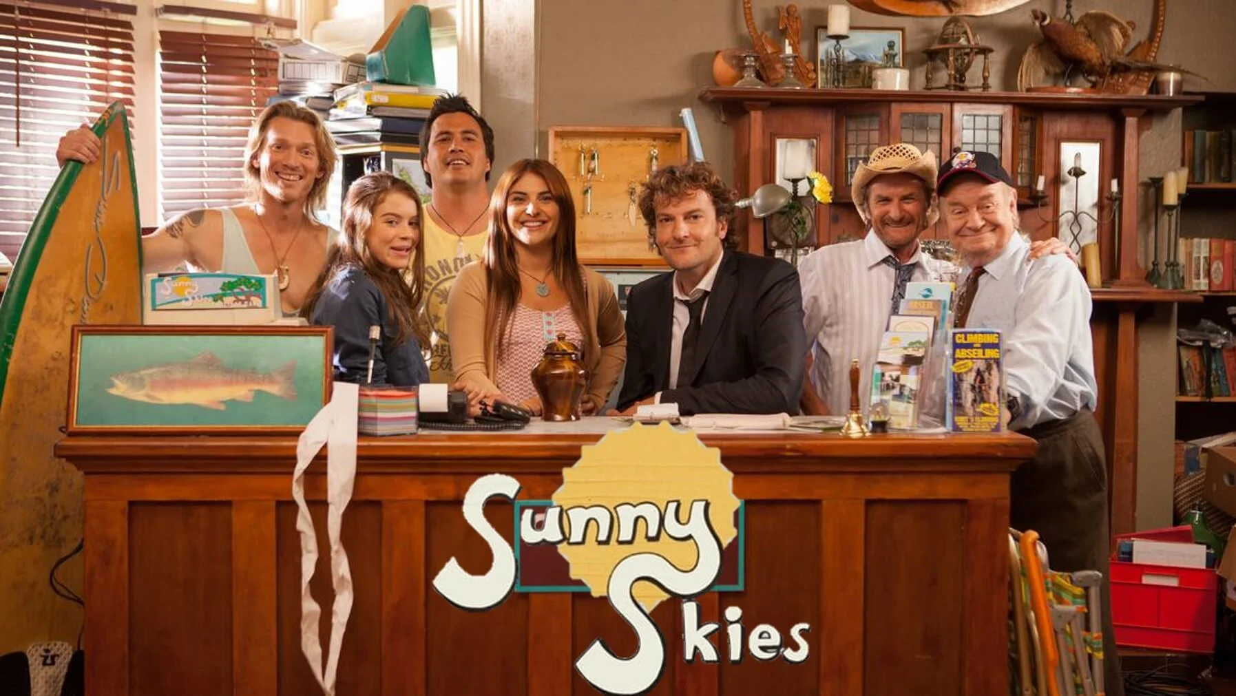 Group of people standing behind a wooden reception desk with the logo "Sunny Skies". The desk has a fish painting and brochures. The background includes a cabinet with books and decorative items.