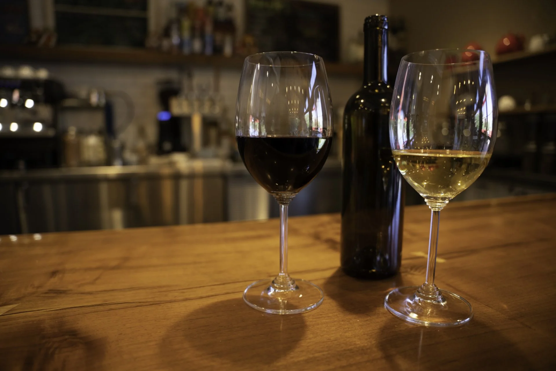 Two wine glasses, one with red wine and one with white wine, placed on a wooden bar counter with a dark bottle in the background.