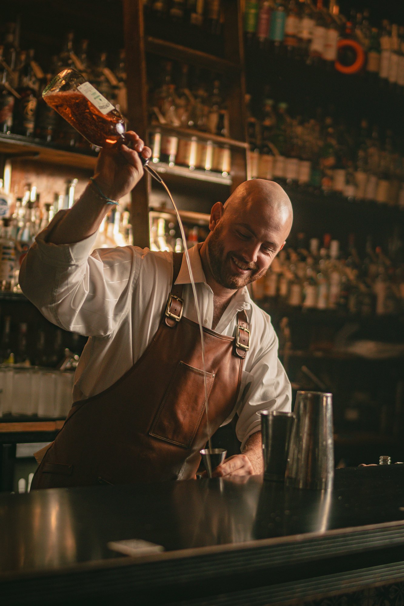 A smiling bartender balancing a bottle of Overeem whiskey on his head in a bar with a shelf full of liquor bottles in the background.