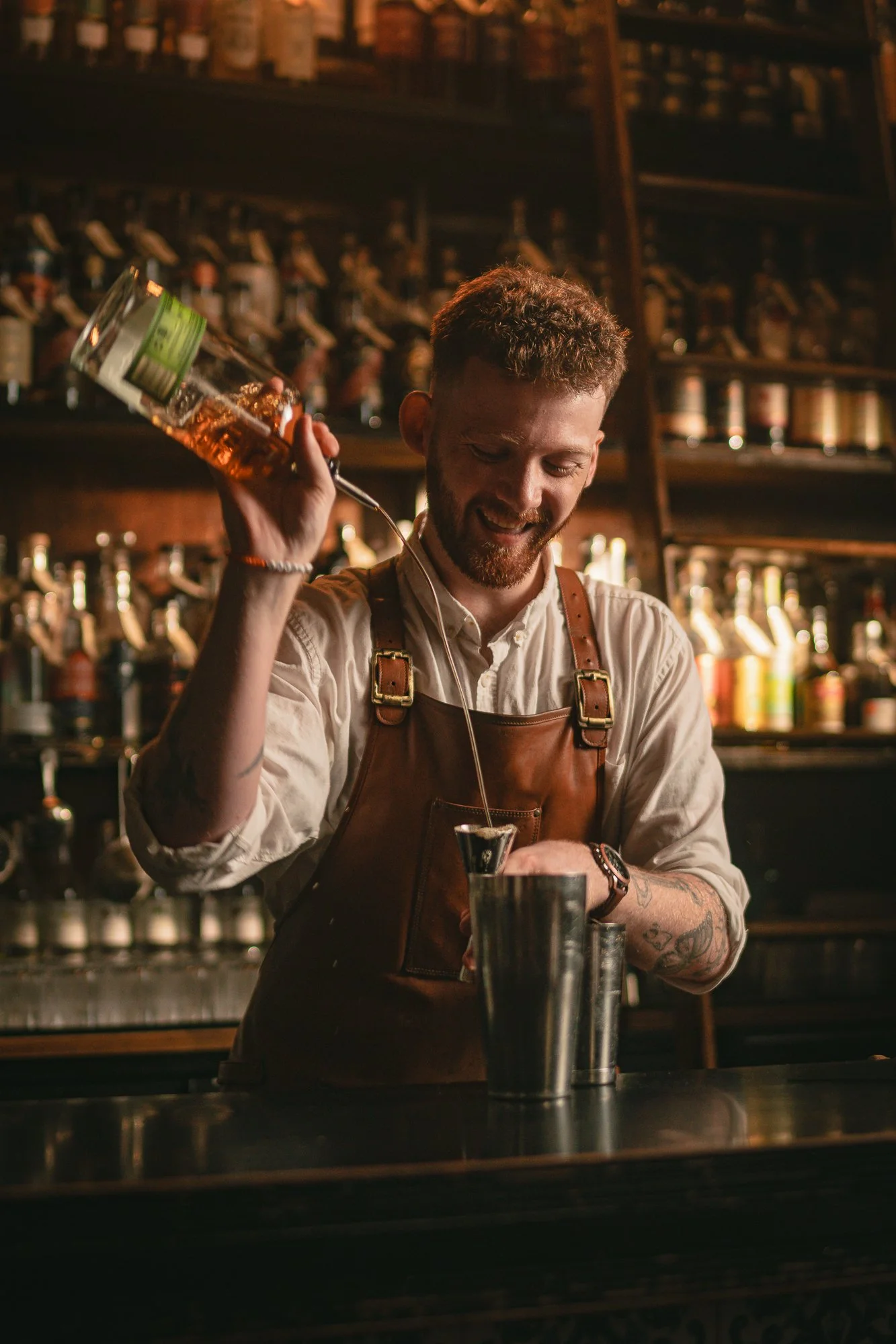 A smiling man with a beard and short hair wearing a white t-shirt and brown apron, standing in front of shelves filled with bottles.