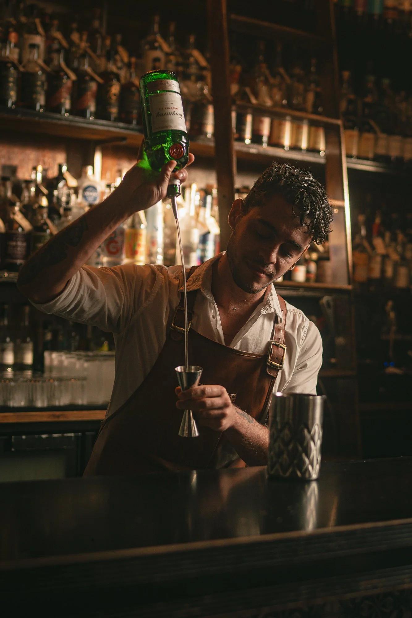 A bartender with tattoos in a white t-shirt and brown apron pouring a drink from a green bottle into a shaker behind a bar.