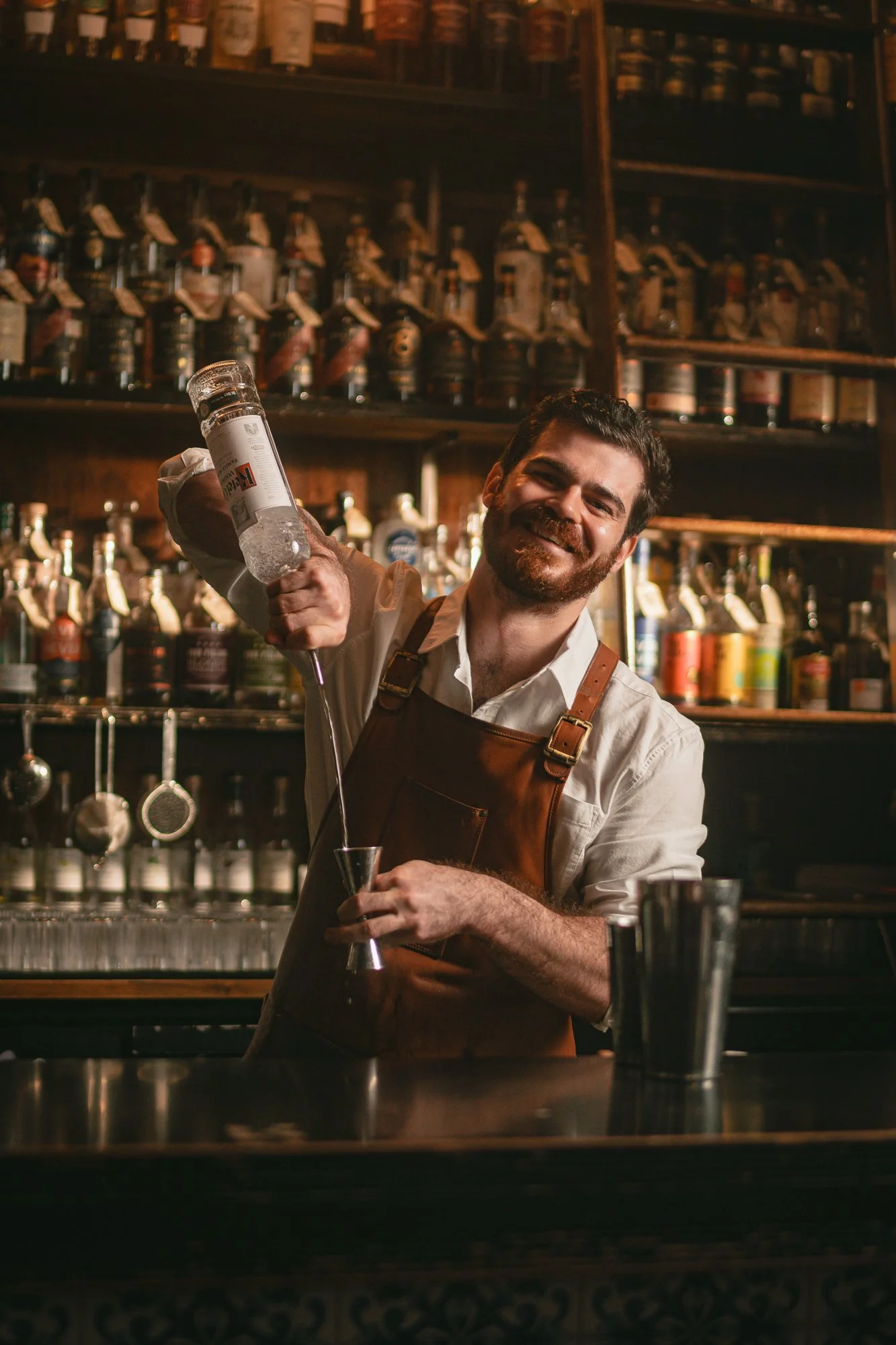 A smiling man in a white t-shirt and brown leather apron standing behind a bar with liquor bottles in the background.