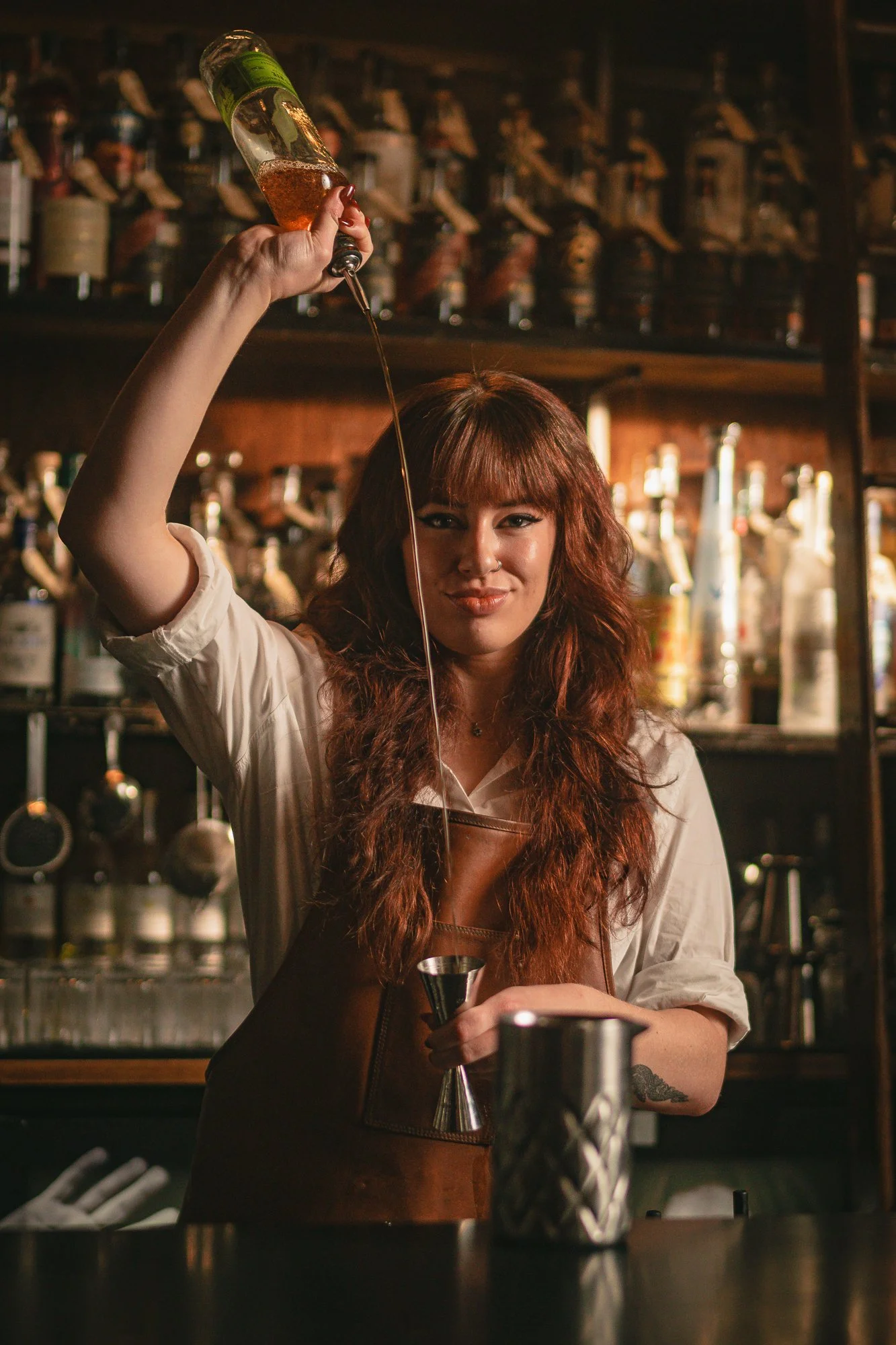 A bartender in a brown apron shakes a cocktail shaker behind a bar with liquor bottles on shelves.