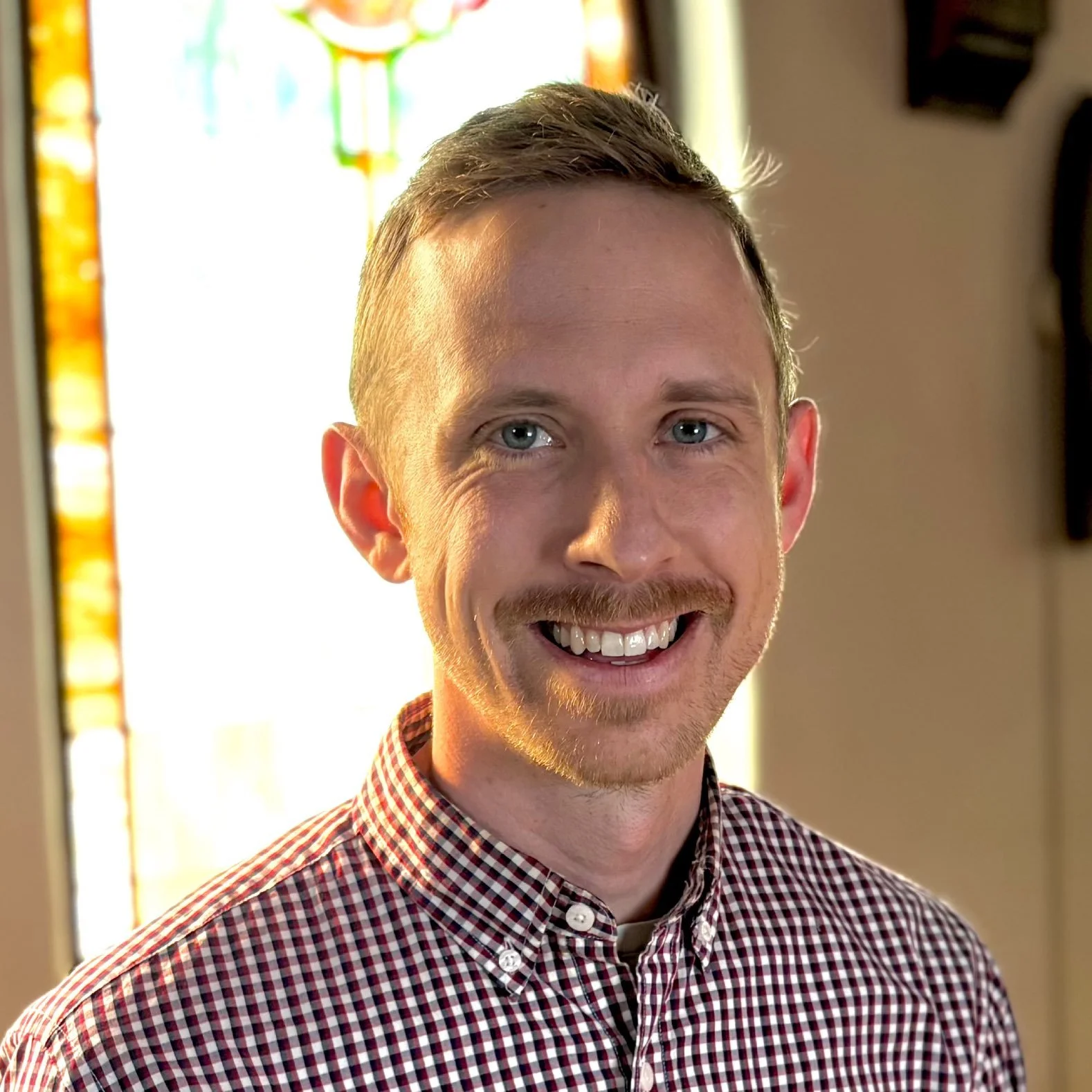 A smiling woman with glasses and short gray hair, wearing a black clerical shirt with a white clerical collar, standing indoors near a colorful stained glass window.