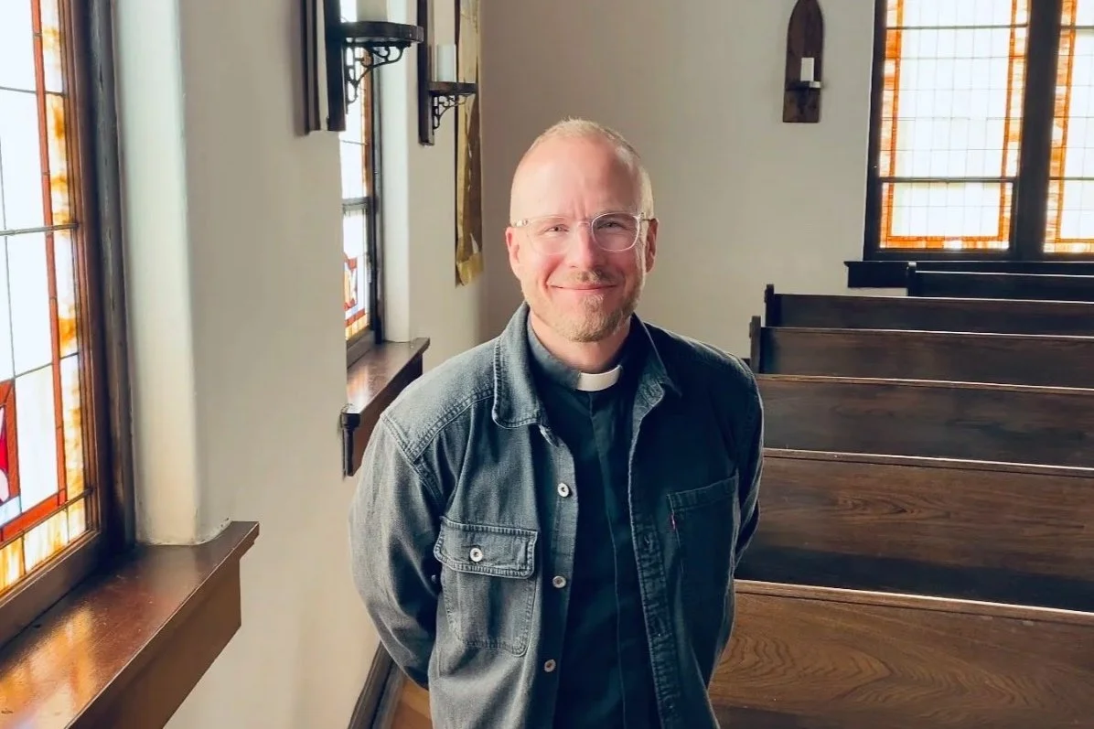 A man with glasses, a beard, and a clerical collar smiling indoors with a softly lit background.
