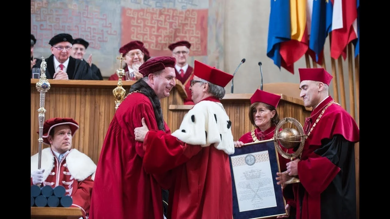 Graduation ceremony with individuals in traditional academic regalia, including robes and caps, in a formal setting with flags in the background and scholarly officials seated at a dais.