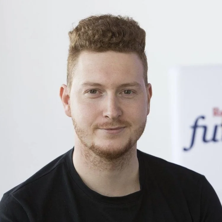 A young man with light skin, reddish curly hair, and a beard, wearing a black shirt, posing for a photo indoors.