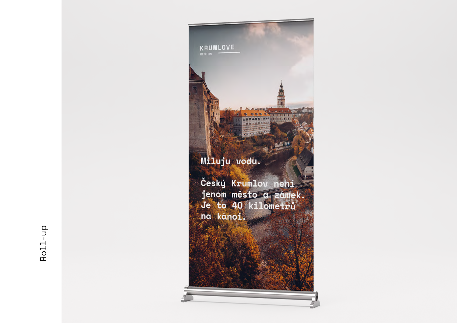 A poster roll-up banner displaying a scenic view of Krumlove, Czech Republic, with historic buildings, a river, and autumn foliage, overlaid with Czech text.