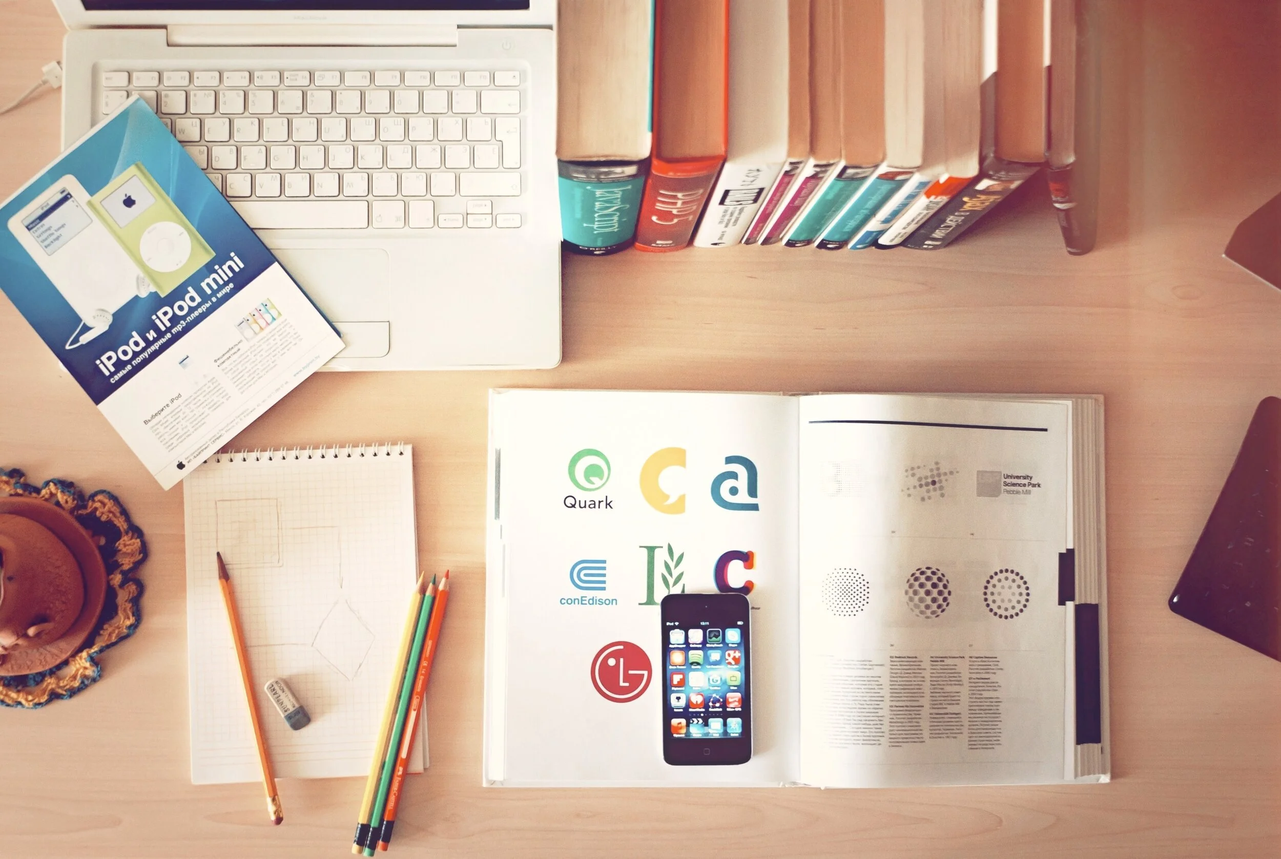 Top-down view of a wooden desk with a white laptop, several books, a flyer about iPod and iPod mini, a spiral notepad with a pencil, colored pencils, a smartphone on an open magazine with tech logos, and a container with cookies.