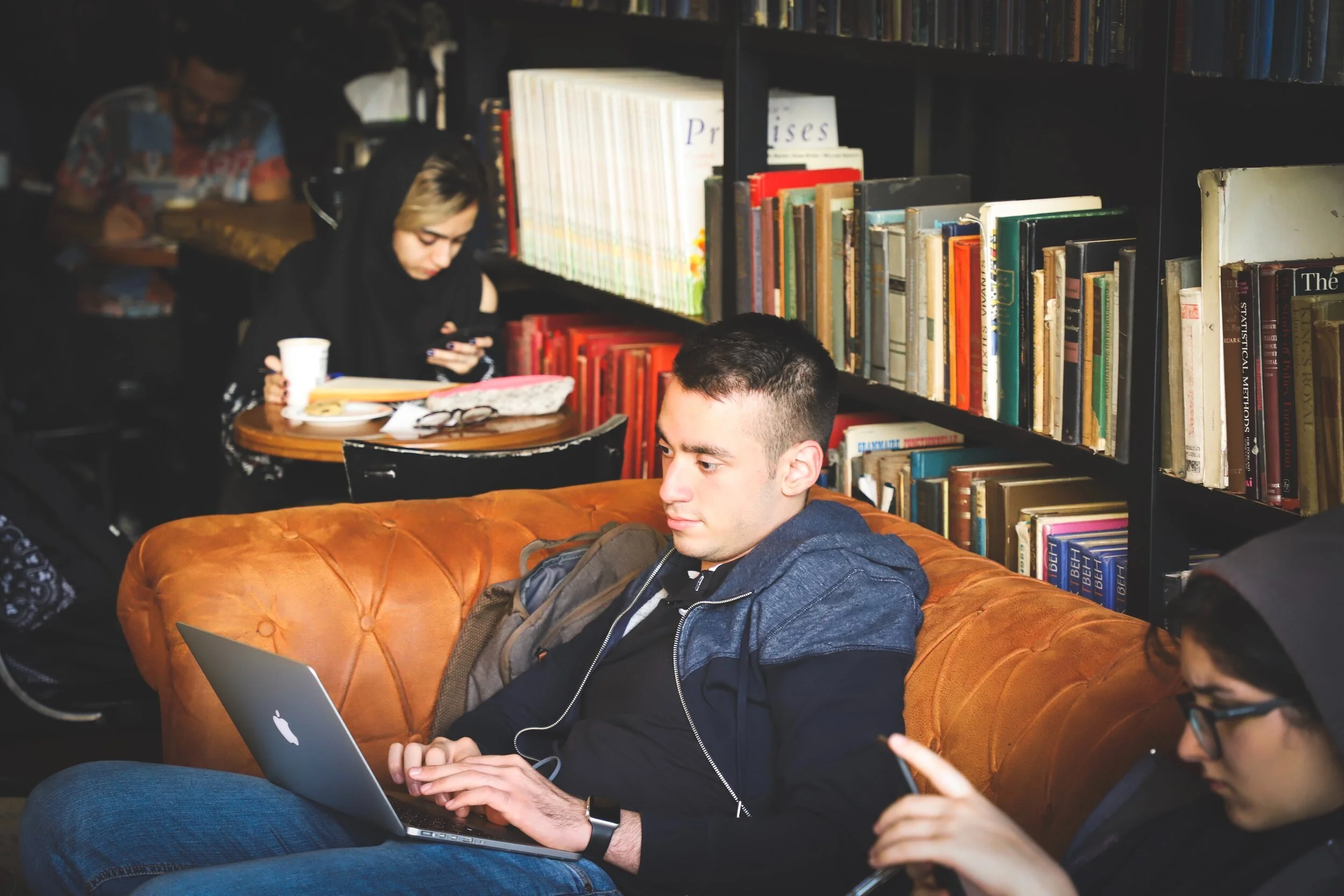 Three young adults in a library or bookstore: a young man sitting on a brown leather couch with a laptop, and two young women sitting at a small round table in the background, one looking at her phone and the other using her phone.