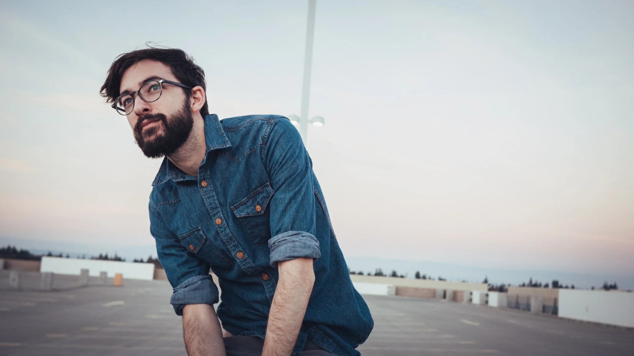 A man with glasses and a beard wearing a denim shirt, looking pensively while leaning forward in an empty parking lot during sunset.