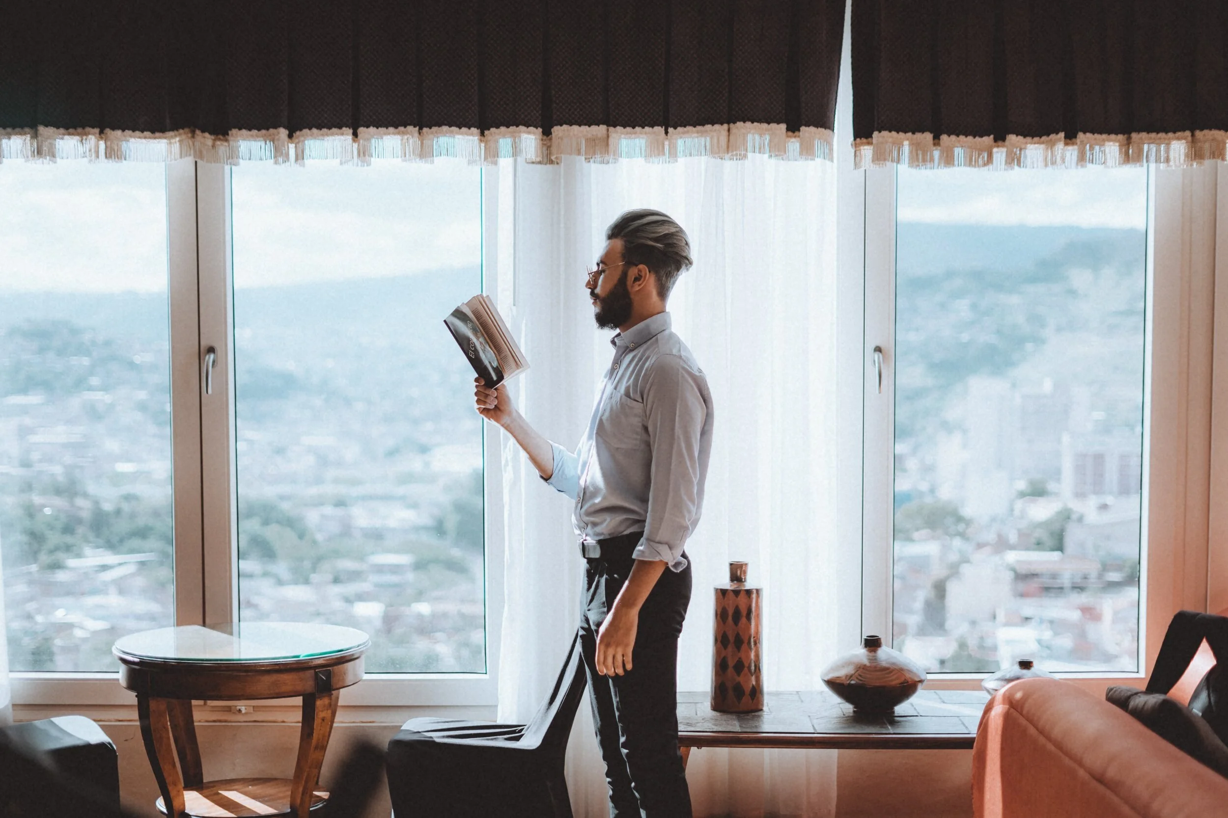 A man with glasses and a beard reads a magazine while standing near large windows in a high-rise apartment. The room has a small wooden table, decorative vases, and a couch.