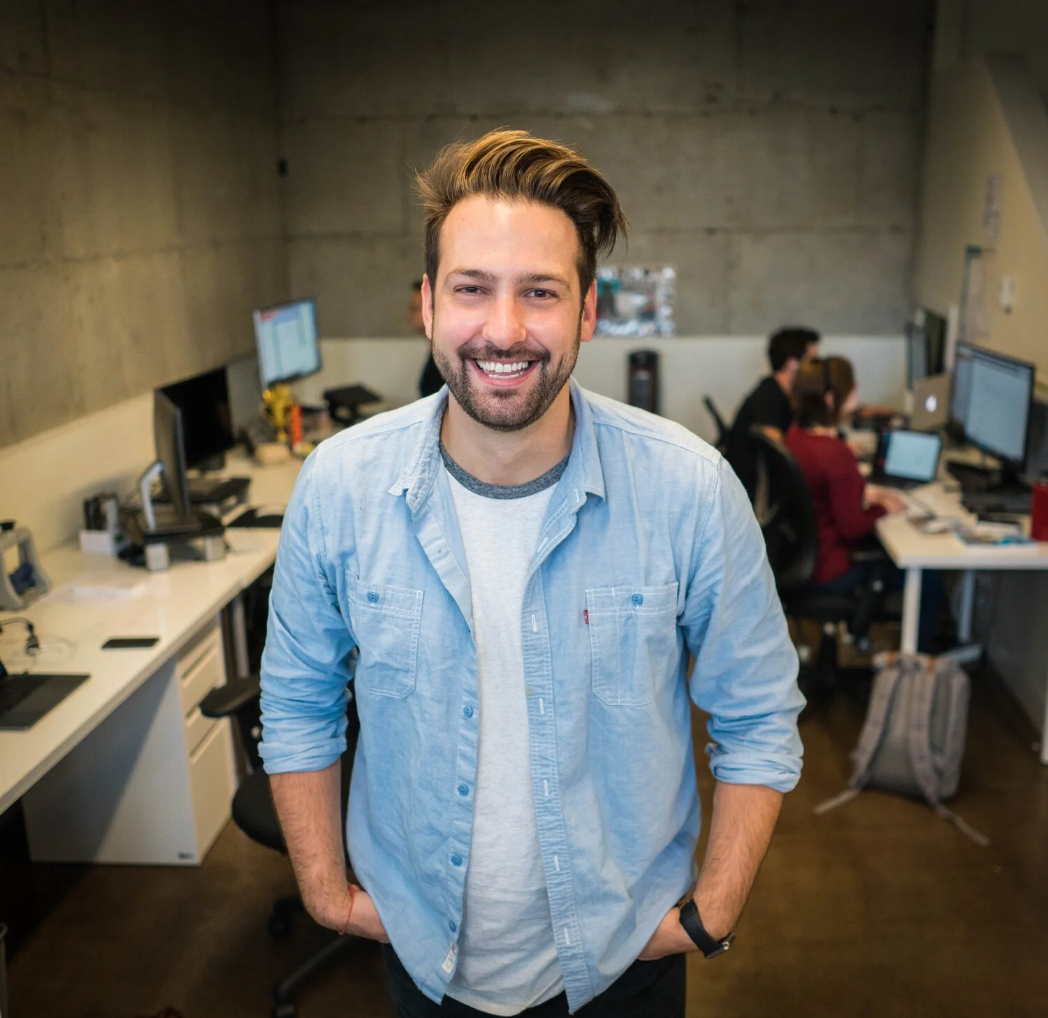 A man with a beard and styled hair smiling and posing in an office with other employees working at desks in the background.