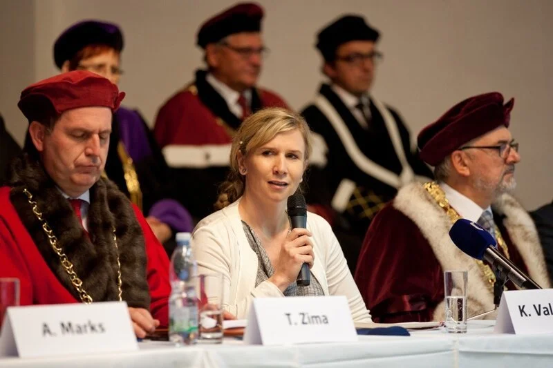 Woman speaking into a microphone at a formal event, surrounded by men in academic regalia with red and purple robes and tassels.