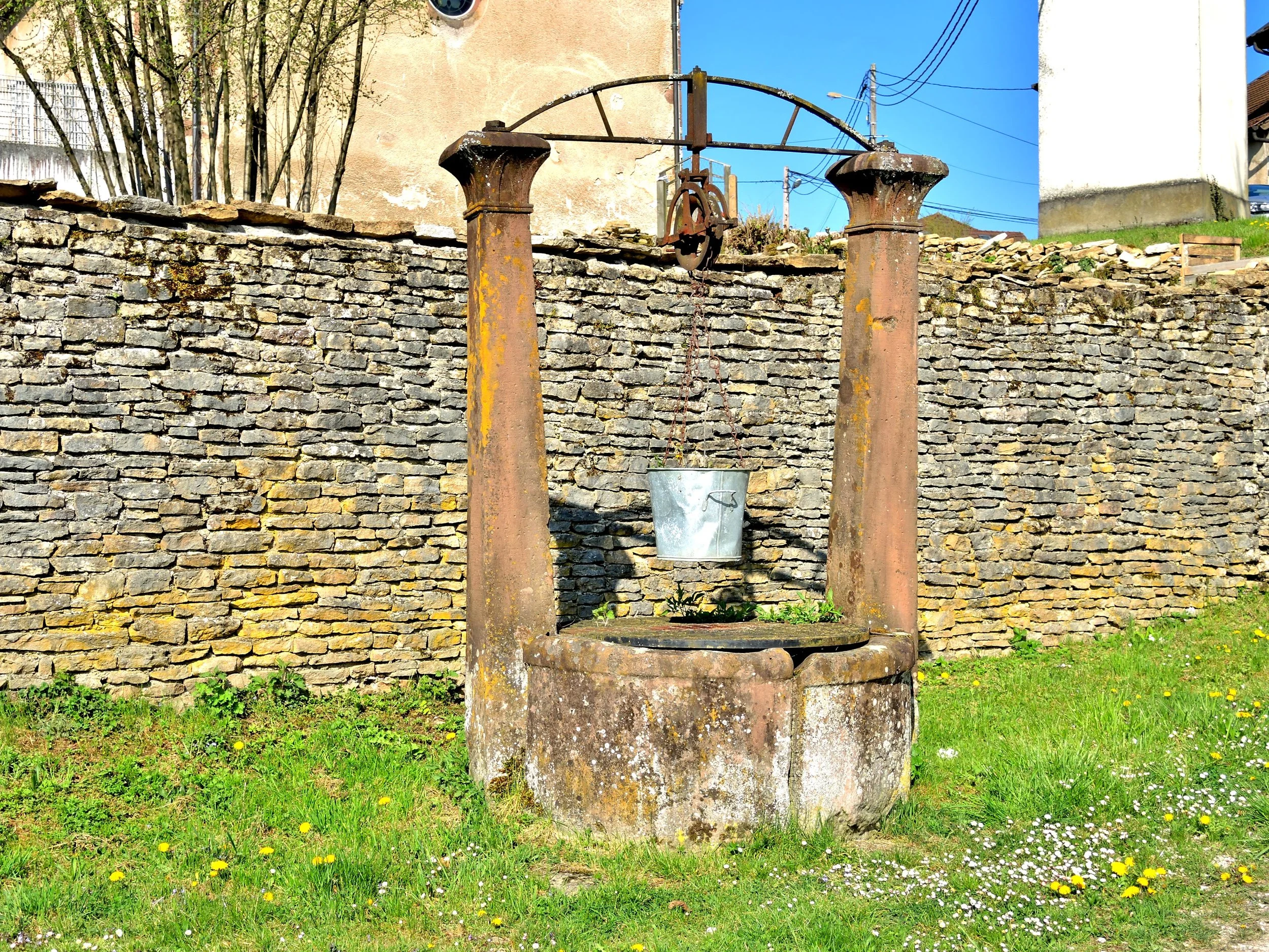 Ancienne auge en pierre avec deux piliers en fer rouillés et un seau suspendu, située dans un jardin avec herbe et petites fleurs, devant un mur en pierre et un bâtiment en arrière-plan, sous un ciel bleu.