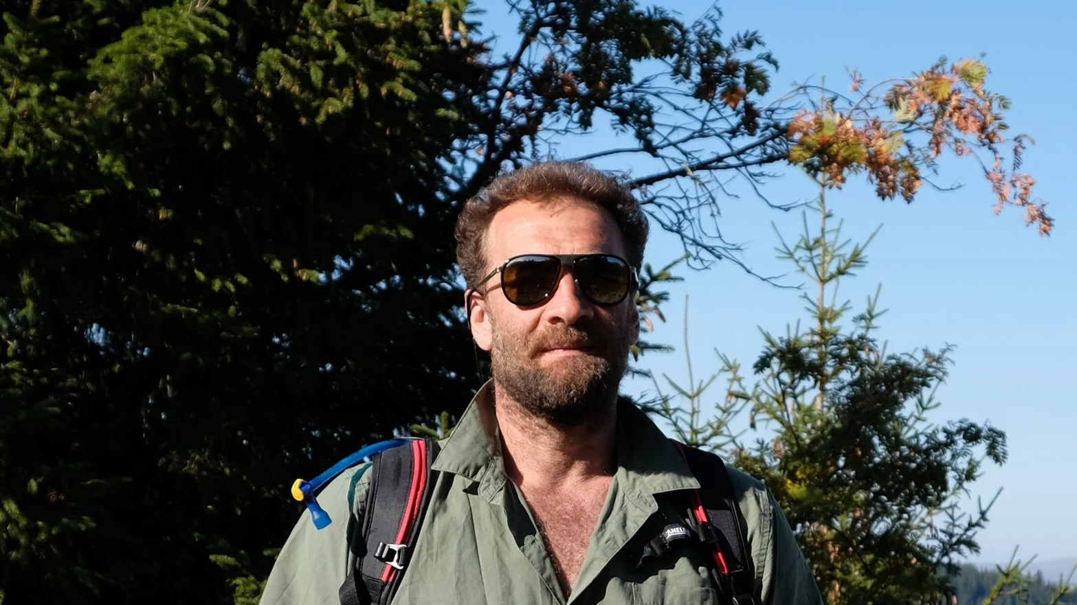 Un homme portant des lunettes de soleil, une chemise verte et un sac à dos, dans un environnement naturel avec des arbres et un ciel bleu.