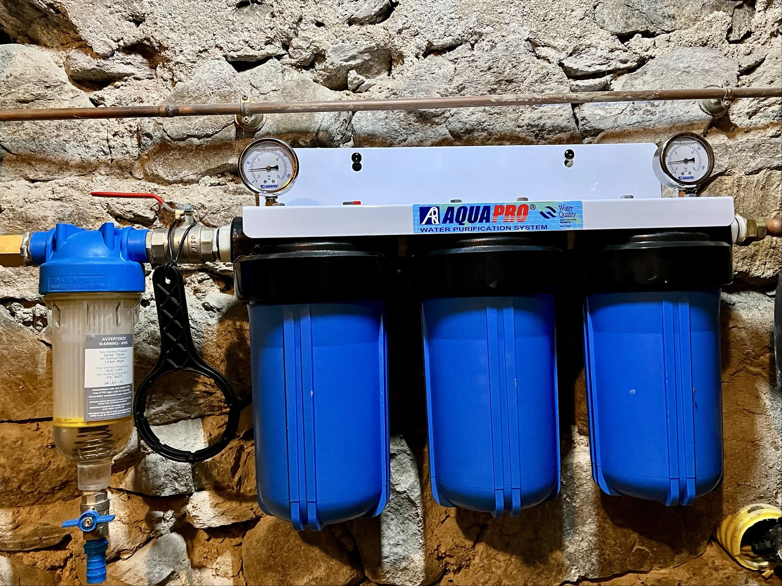 Filtre de purification d'eau avec trois cartouches bleues, accessoires en cuivre, manomètres et un tuyau noir, installé contre une vieille pierre.