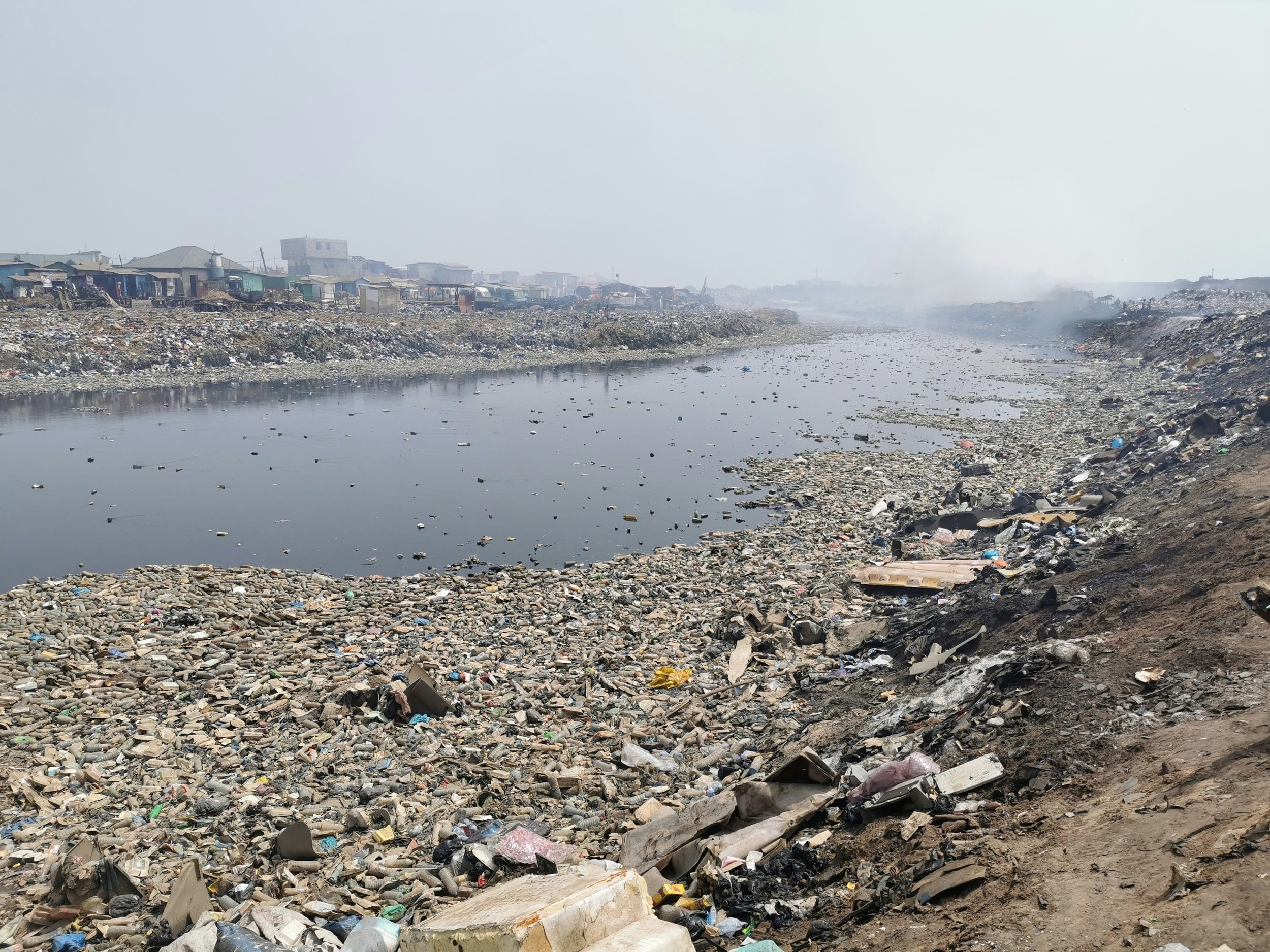 Un rivage sale avec beaucoup de déchets et de débris plastiques, une rivière polluée et des habitations en arrière-plan, atmosphère brumeuse ou polluée.
