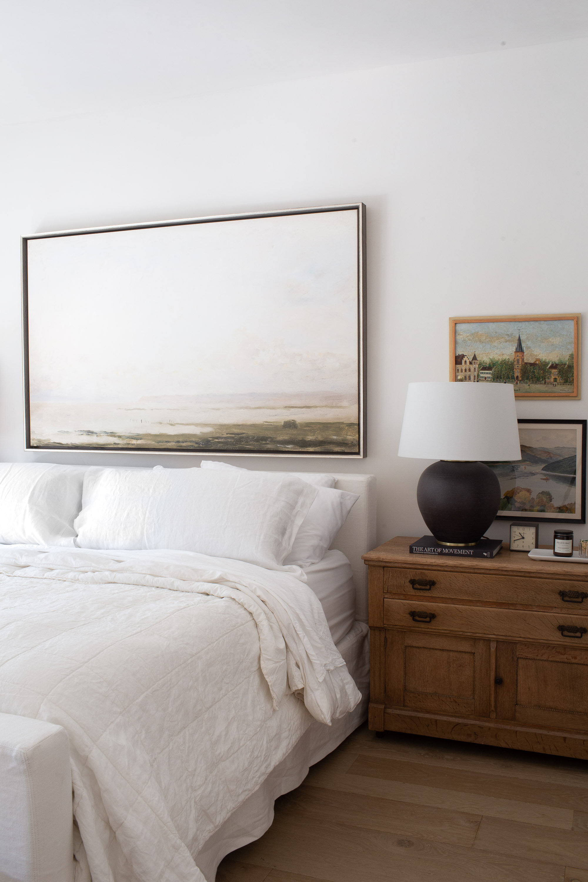 View of a side of bed covered in white linen with an antique side dresser and landscape scene hanging above the bed.