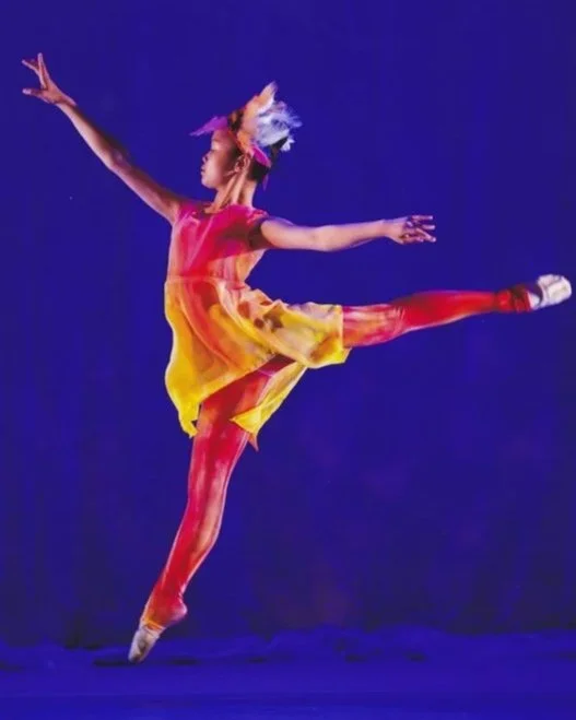 A young girl in a colorful dress and feathered headband performs a ballet leap against a blue background.