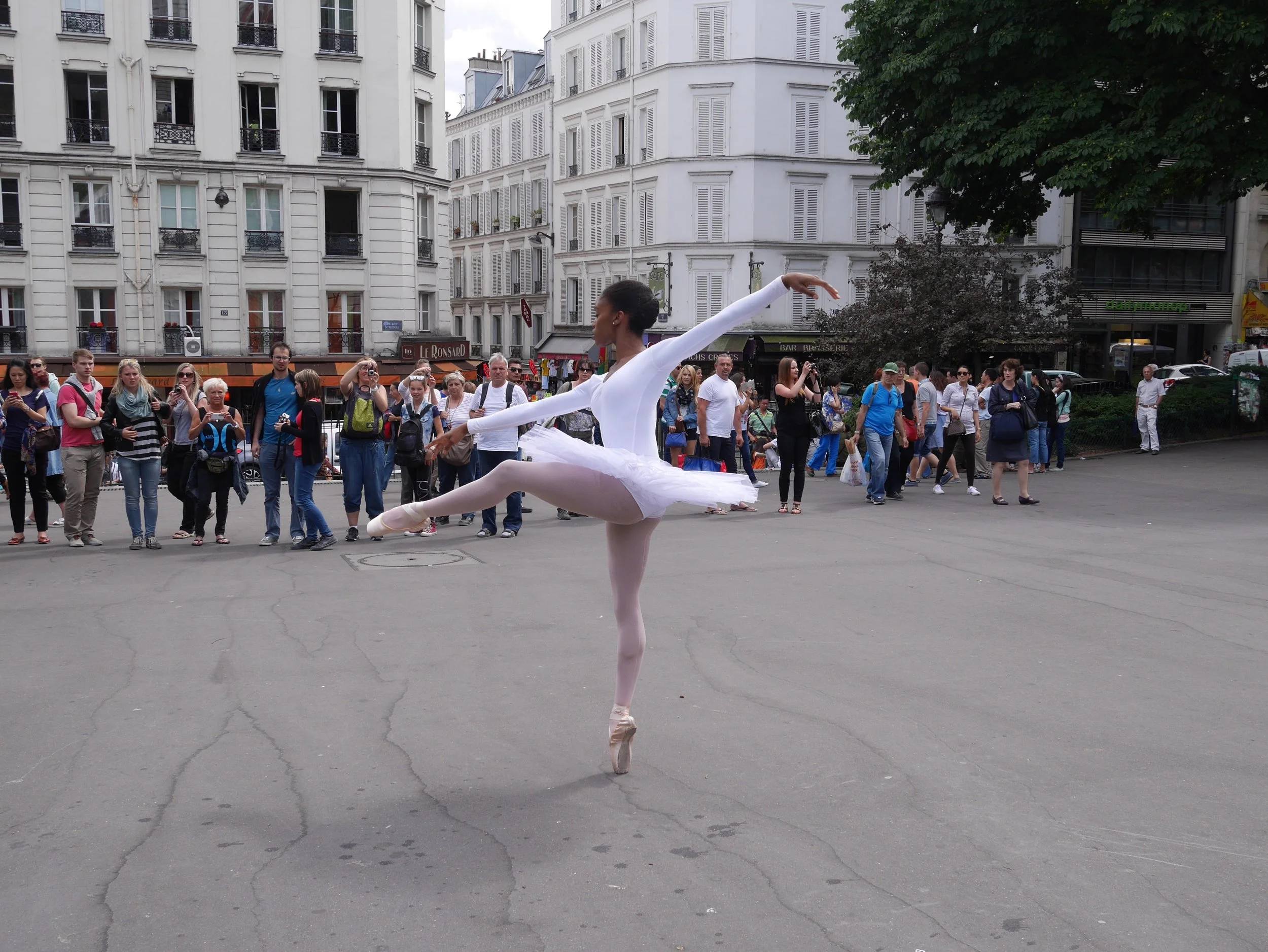 A ballet dancer performing a pirouette on an outdoor city street with a crowd of spectators and tall white buildings in the background.