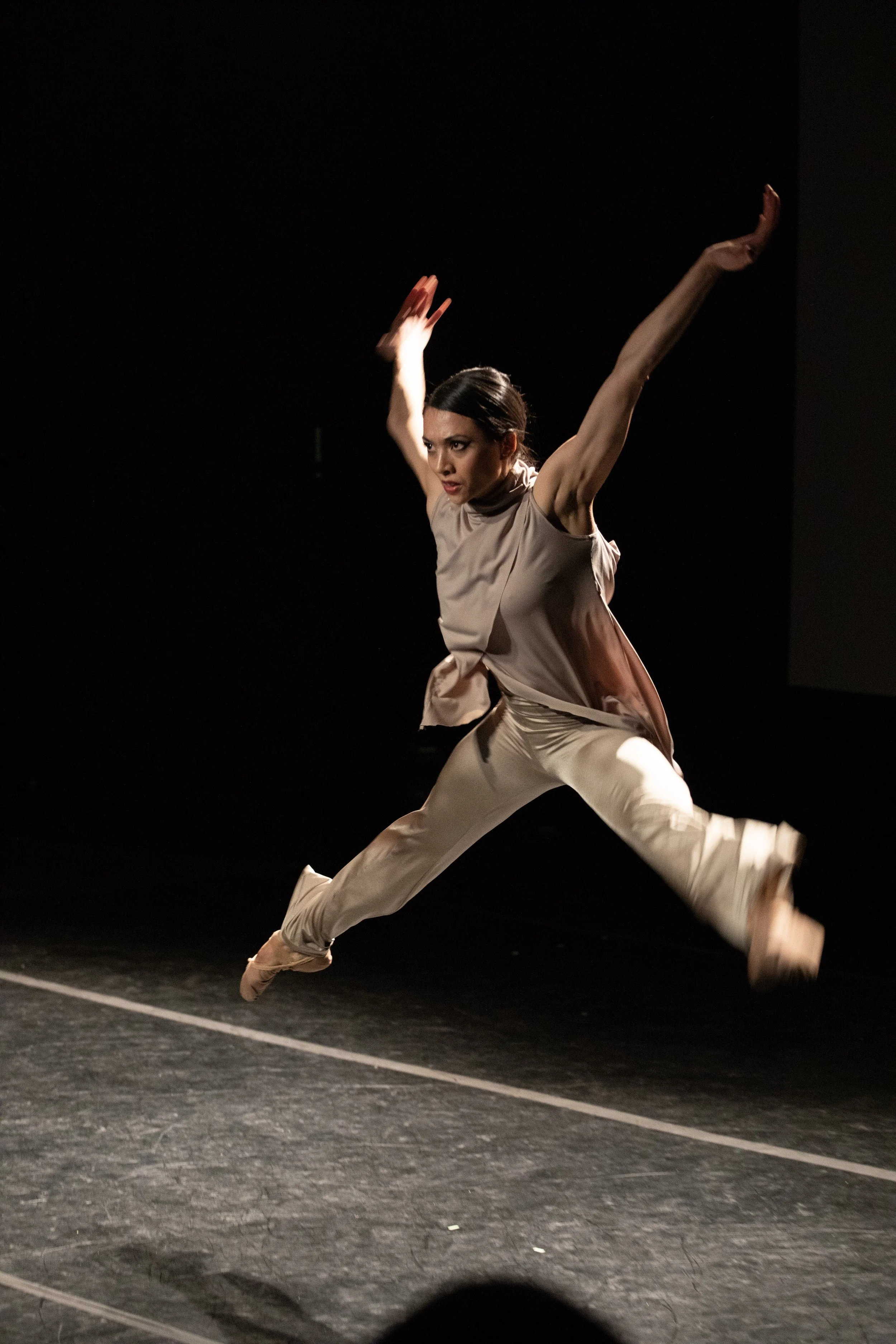 A female dancer in beige clothing performing an energetic jump on a dark stage, with arms raised and legs extended.