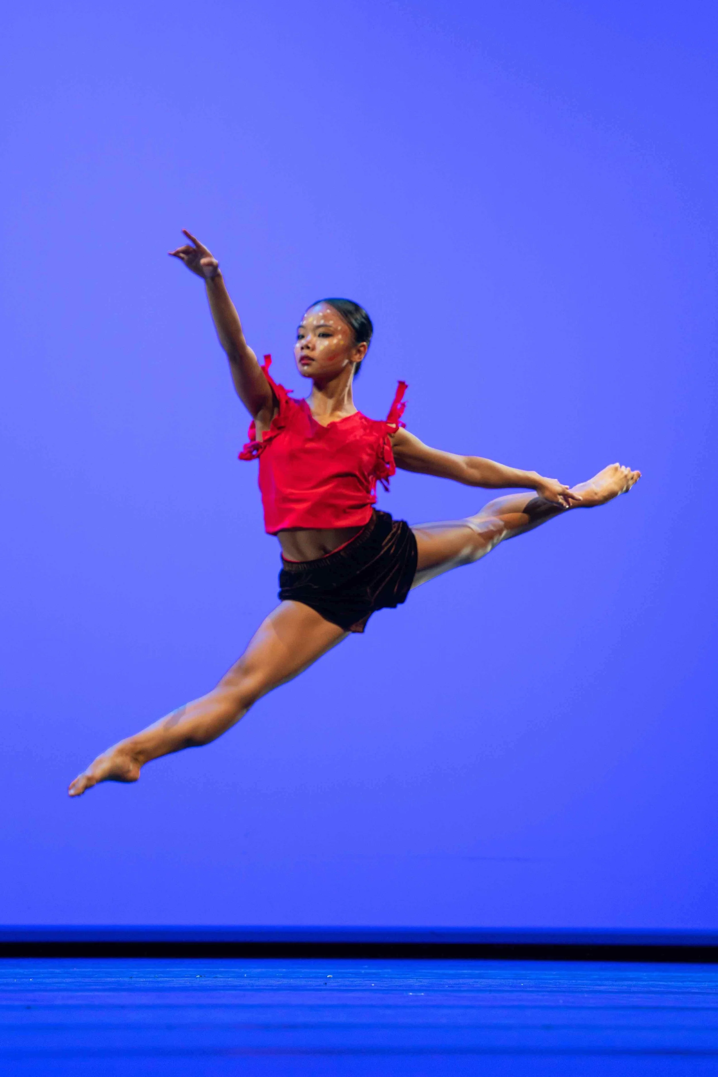 A female dancer performing an acrobatic leap on stage with a solid blue background.