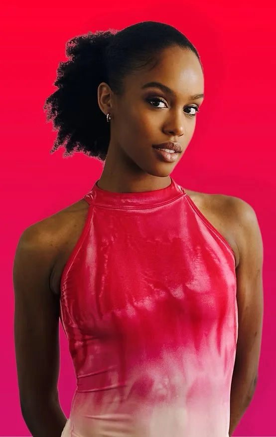 A young woman with dark skin, curly black hair, and wearing earrings, poses against a vibrant pink background. She is dressed in a shiny red sleeveless top.