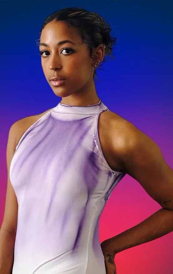 A young woman with dark curly hair in a short style, wearing a shiny lavender sleeveless top, posing against a vibrant gradient background of blue, purple, and pink.