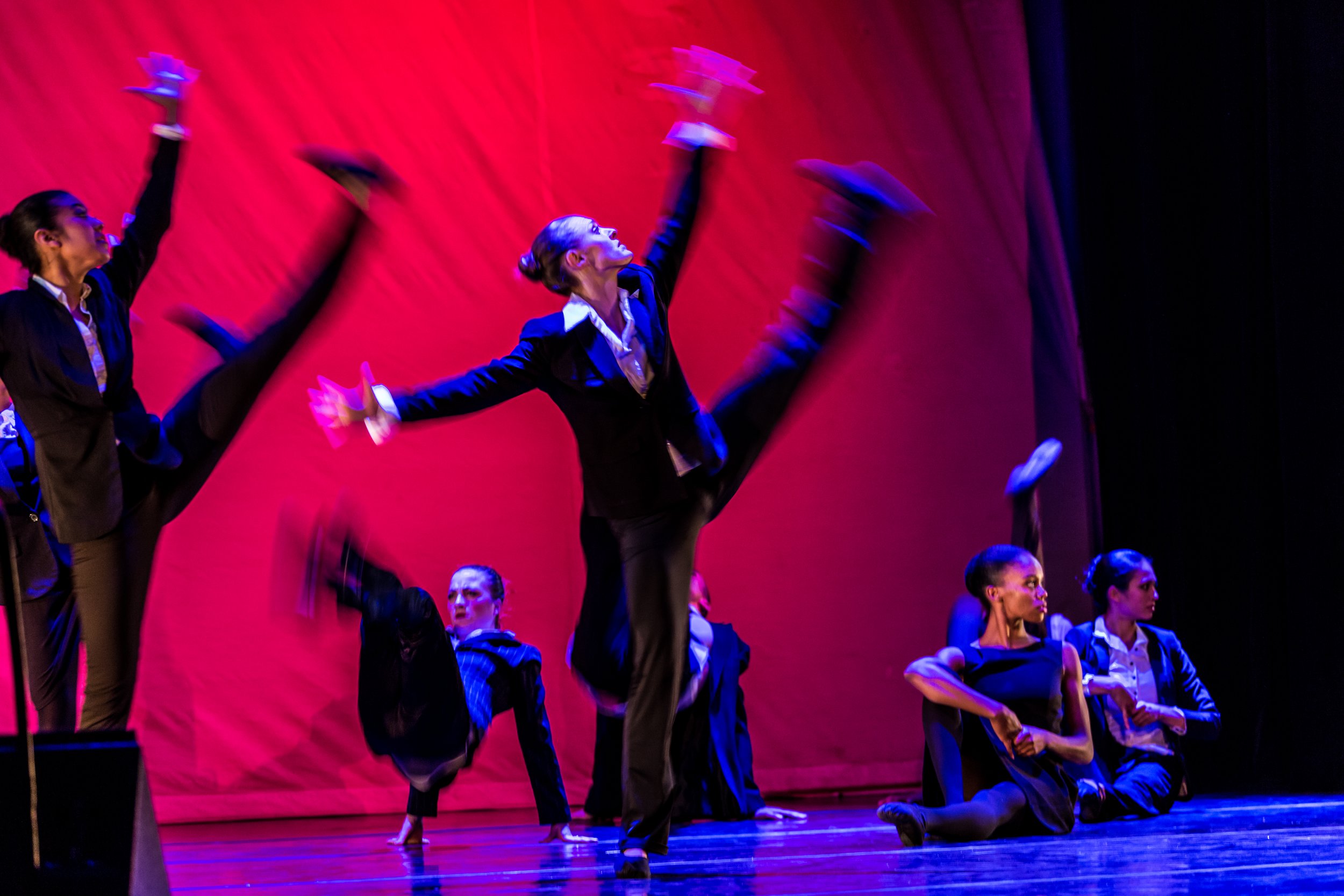 A group of dancers in formal attire performing a ballet on stage with a red backdrop, some dancers are in a kneeling position while others are standing with arms raised.