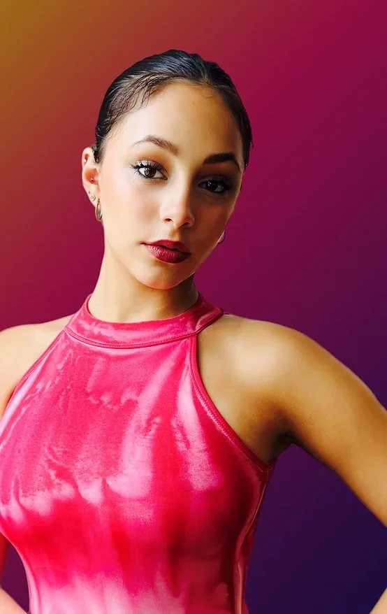 A woman with dark hair styled back, wearing a shiny pink sleeveless top, and looking at the camera with a serious expression, set against a colorful gradient background.