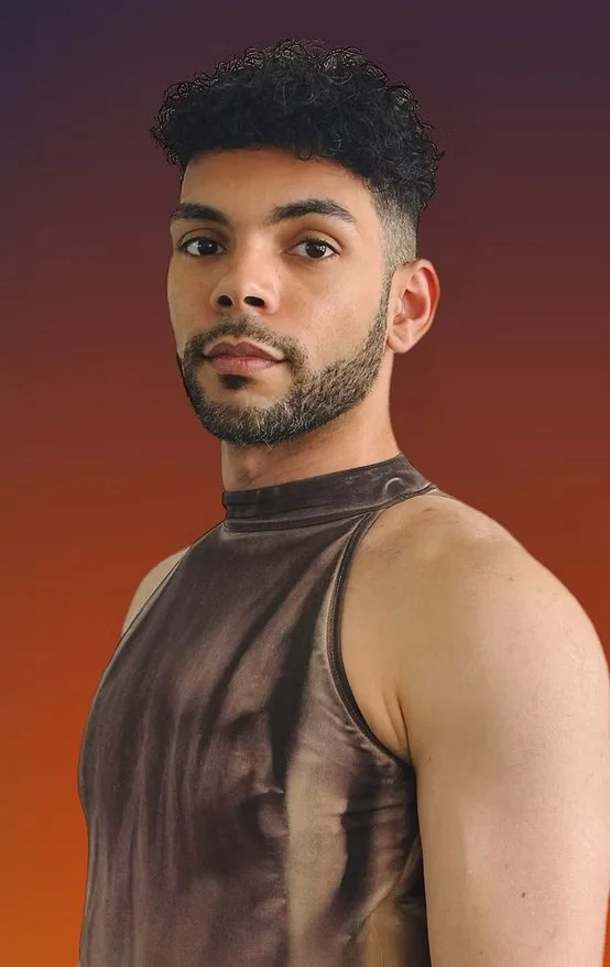 A man with curly black hair and a beard is posing against a gradient background that transitions from purple to orange. He is wearing a sleeveless, shiny, dark-colored top.