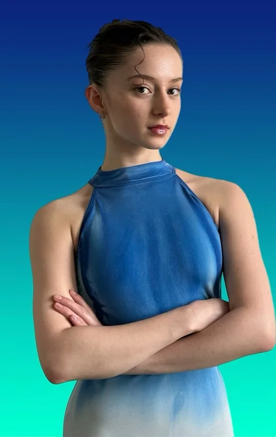 A young woman with hair in a bun, wearing a sleeveless blue dress, posing with crossed arms against a gradient blue background.