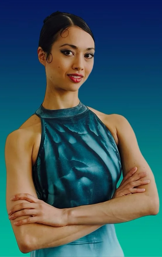 A young woman with dark hair styled in an updo, wearing a sleeveless, shiny blue-green dress, standing with crossed arms against a gradient blue background.