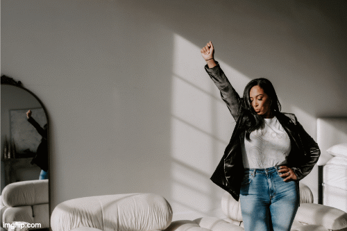 A young woman in a white shirt and jeans, wearing a black leather jacket, standing in a room with sunlight casting shadows from window blinds, raising her right fist in the air in a confident pose.
