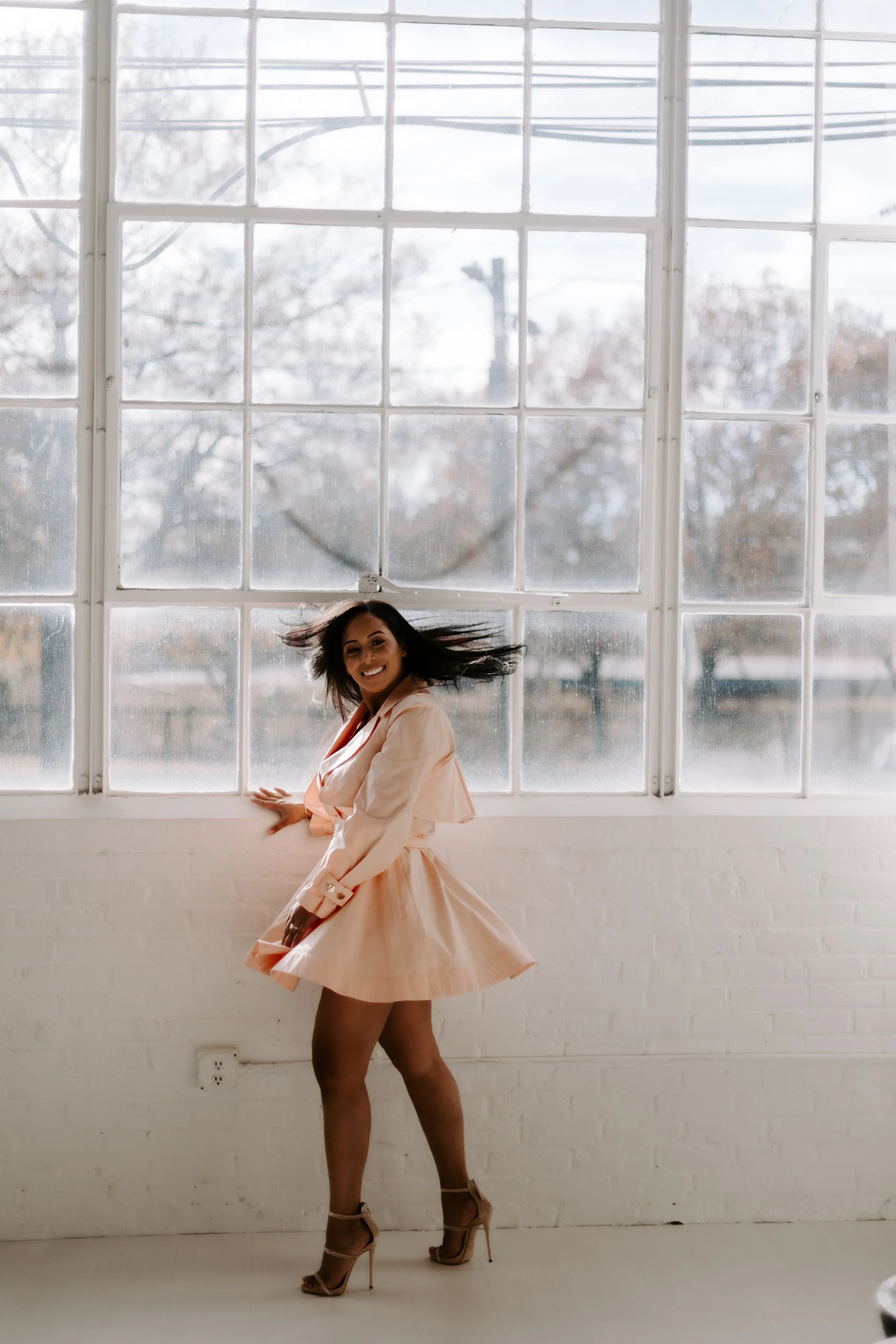 A woman in a peach-colored dress and high heels is dancing or spinning in front of large windows, with trees visible outside and her hair flying.