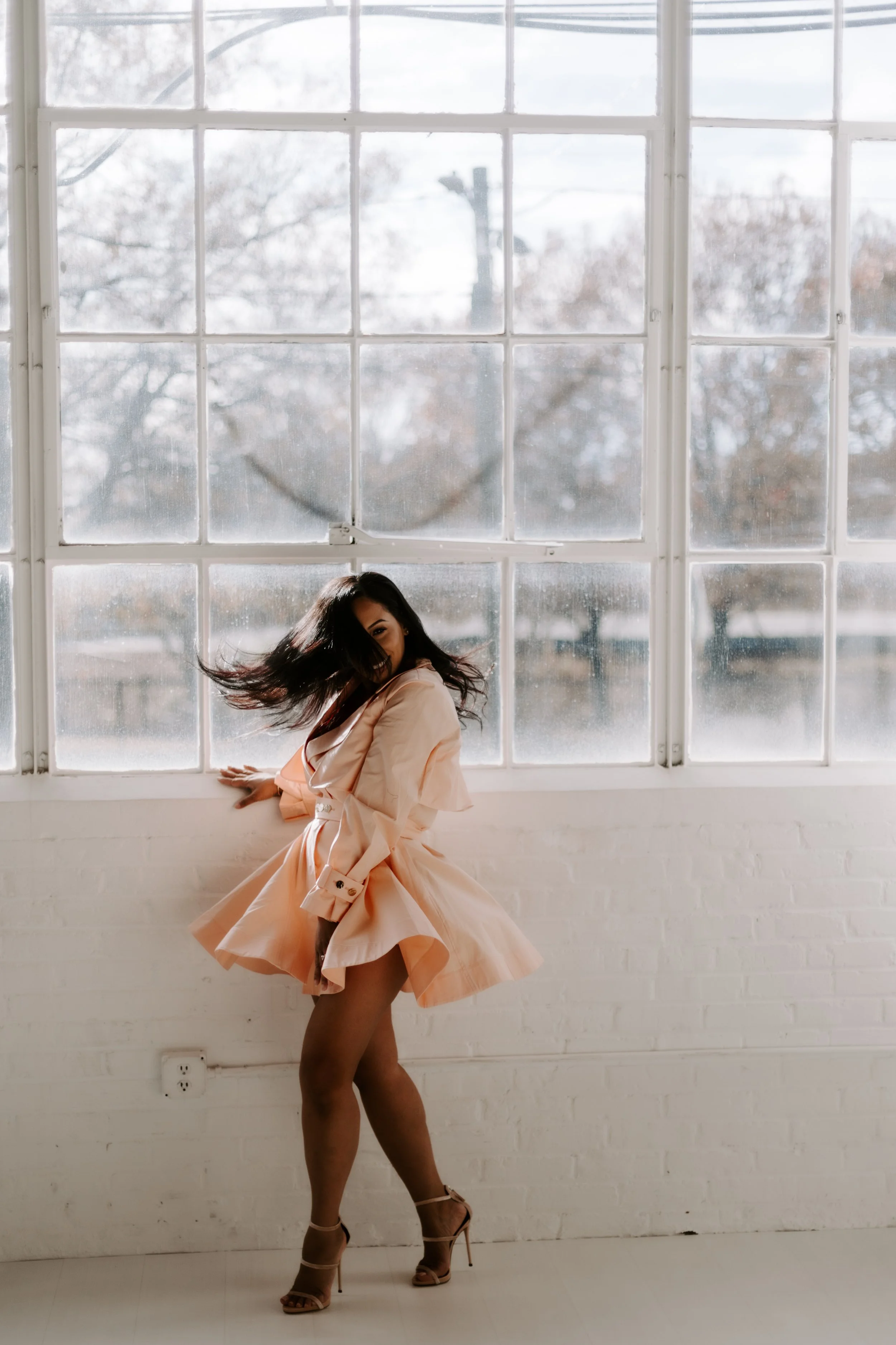 A woman in a peach dress with ruffles and high heels dancing in front of a large window with a white brick wall behind her.