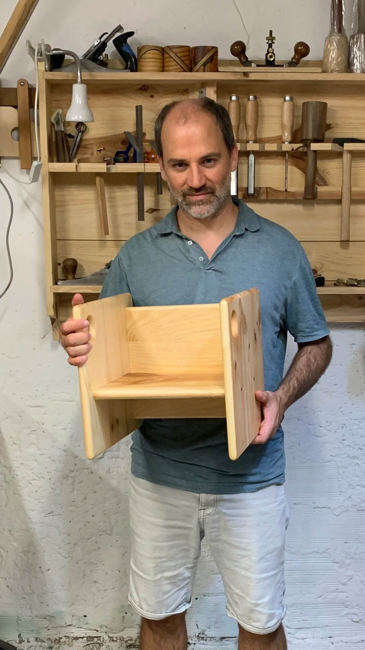 Hombre sosteniendo una silla para niños de tres posturas de madera en un taller de carpintería, con herramientas en una estantería de madera al fondo.