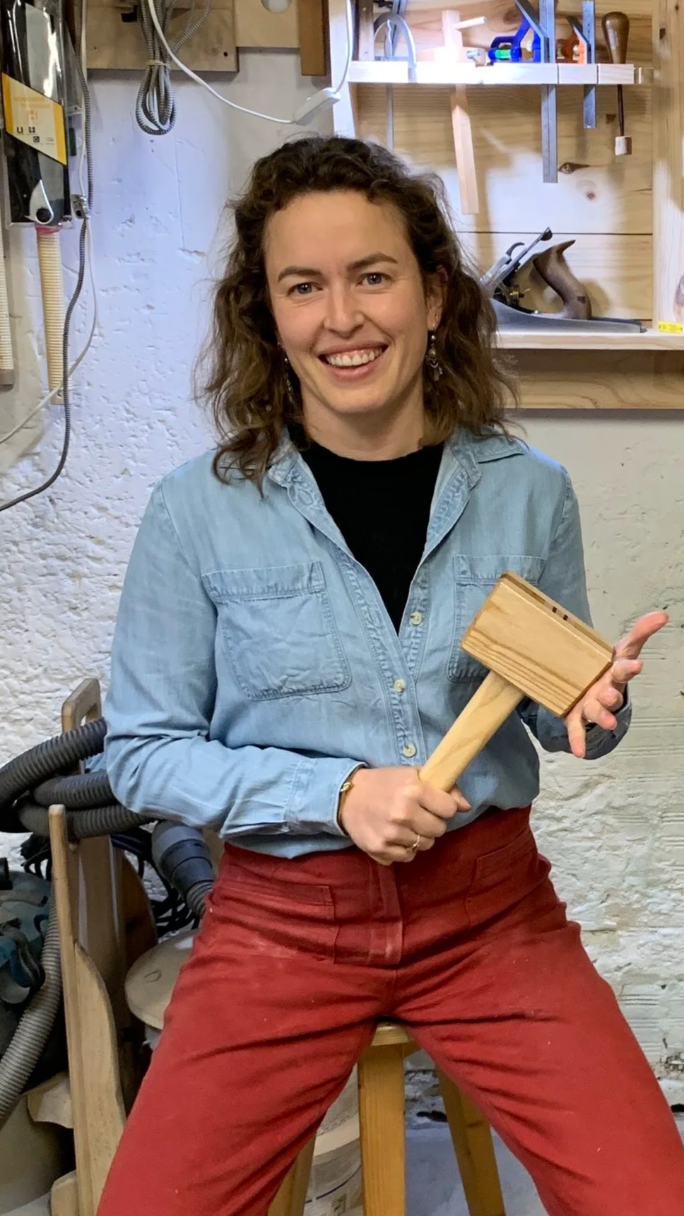 Una mujer sosteniendo una maza de madera en un taller de carpintería, sonriendo.