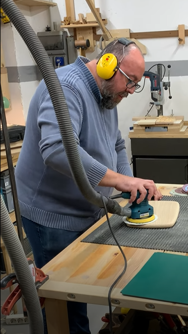 Hombre usando una lijadora eléctrica en un taller de carpintería mientras fabrica una tabla de quesos, con protección auditiva amarilla y gafas.