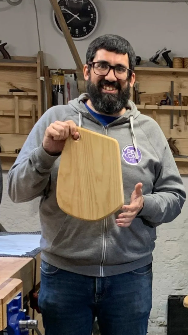 Hombre con barba y sonriendo, gafas y sudadera gris sostiene una tabla para quesos de madera recientemente terminada luego de un curso en un taller de carpintería.