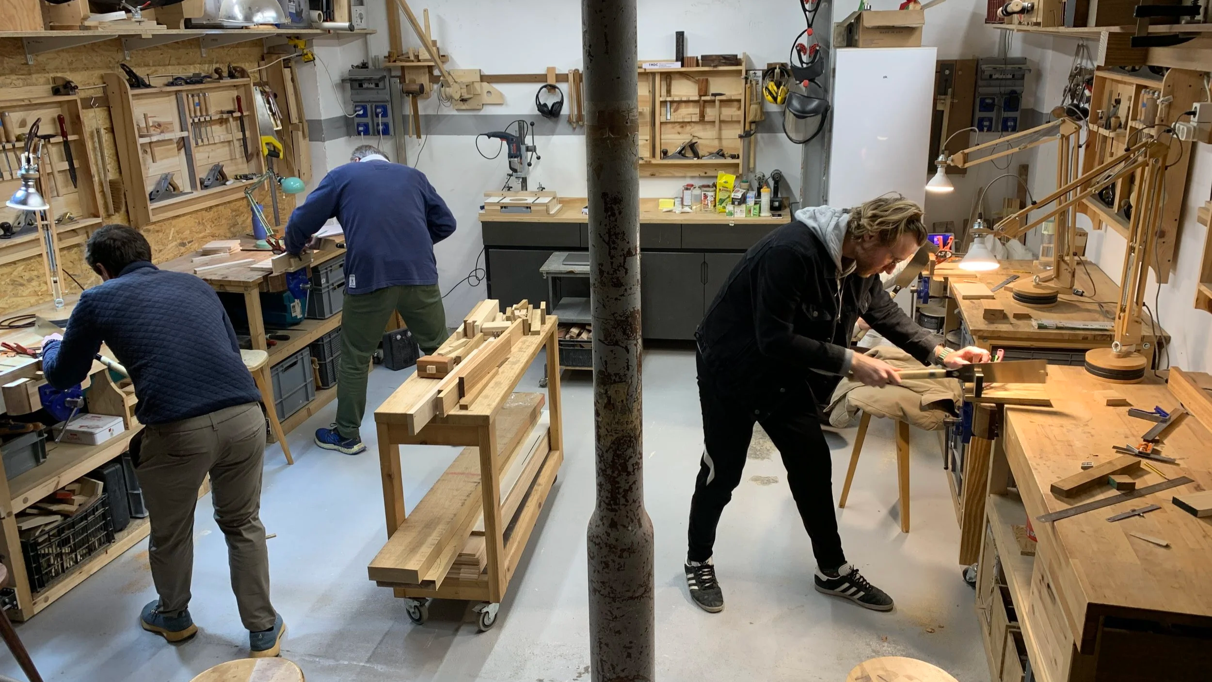 Cuatro personas trabajando en un taller de carpintería, cortando madera y realizando tareas con herramientas. El taller está equipado con mesas de trabajo, estanterías con herramientas y materiales varios.