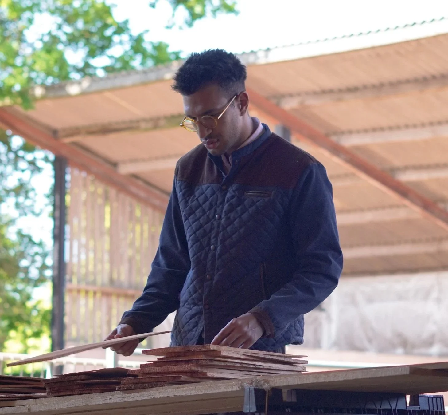 A man with short, curly hair and glasses stand outdoors working on a woodworking project, with a stack of wooden boards on a table in front of him. There are trees and a building structure in the background.