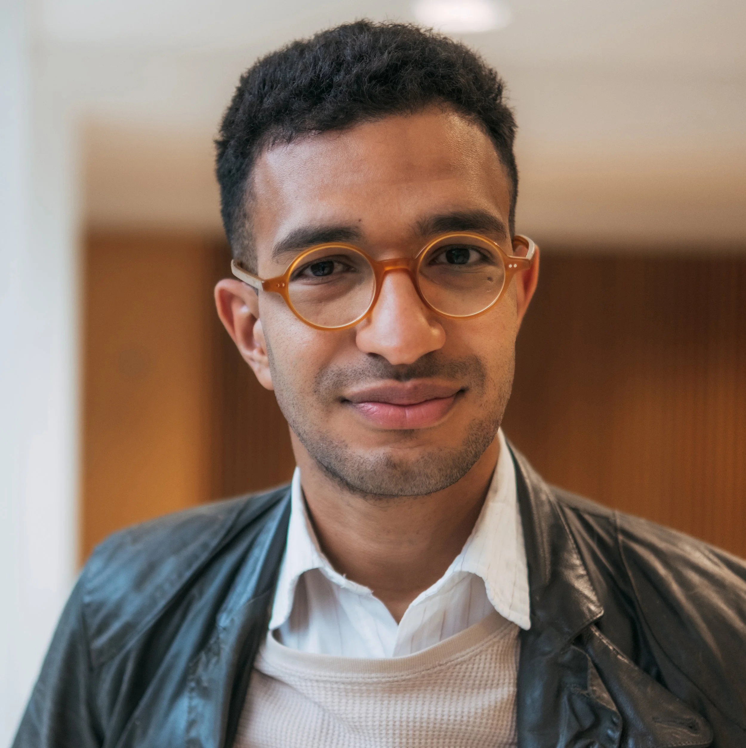 Close-up portrait of Peter Markos with dark curly hair, wearing tan round glasses, a white shirt, and a leather jacket, smiling slightly.