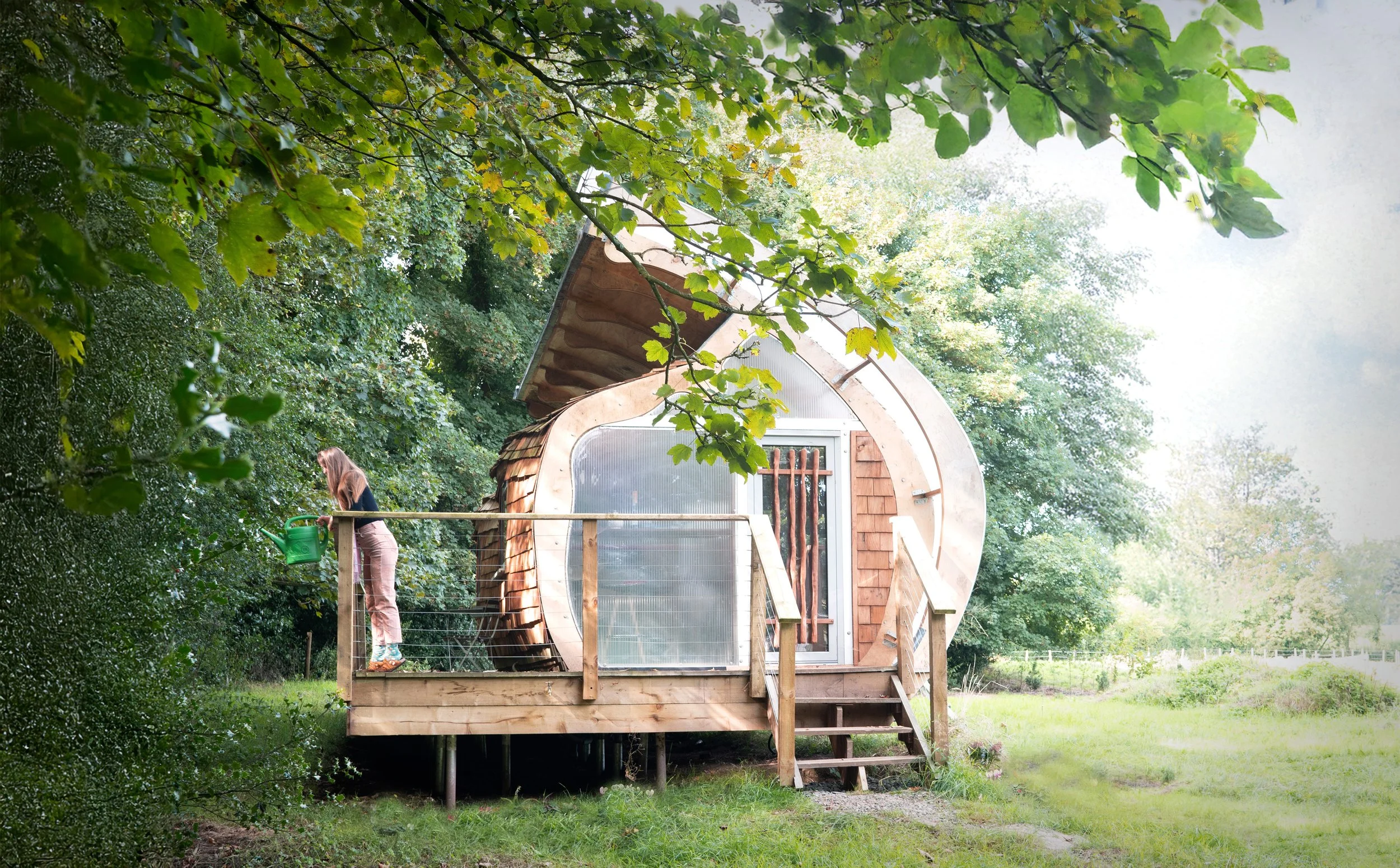 A woman watering plants on a raised wooden deck in front of a small, round, wooden tiny house with a curved roof, set amid green trees and grass.
