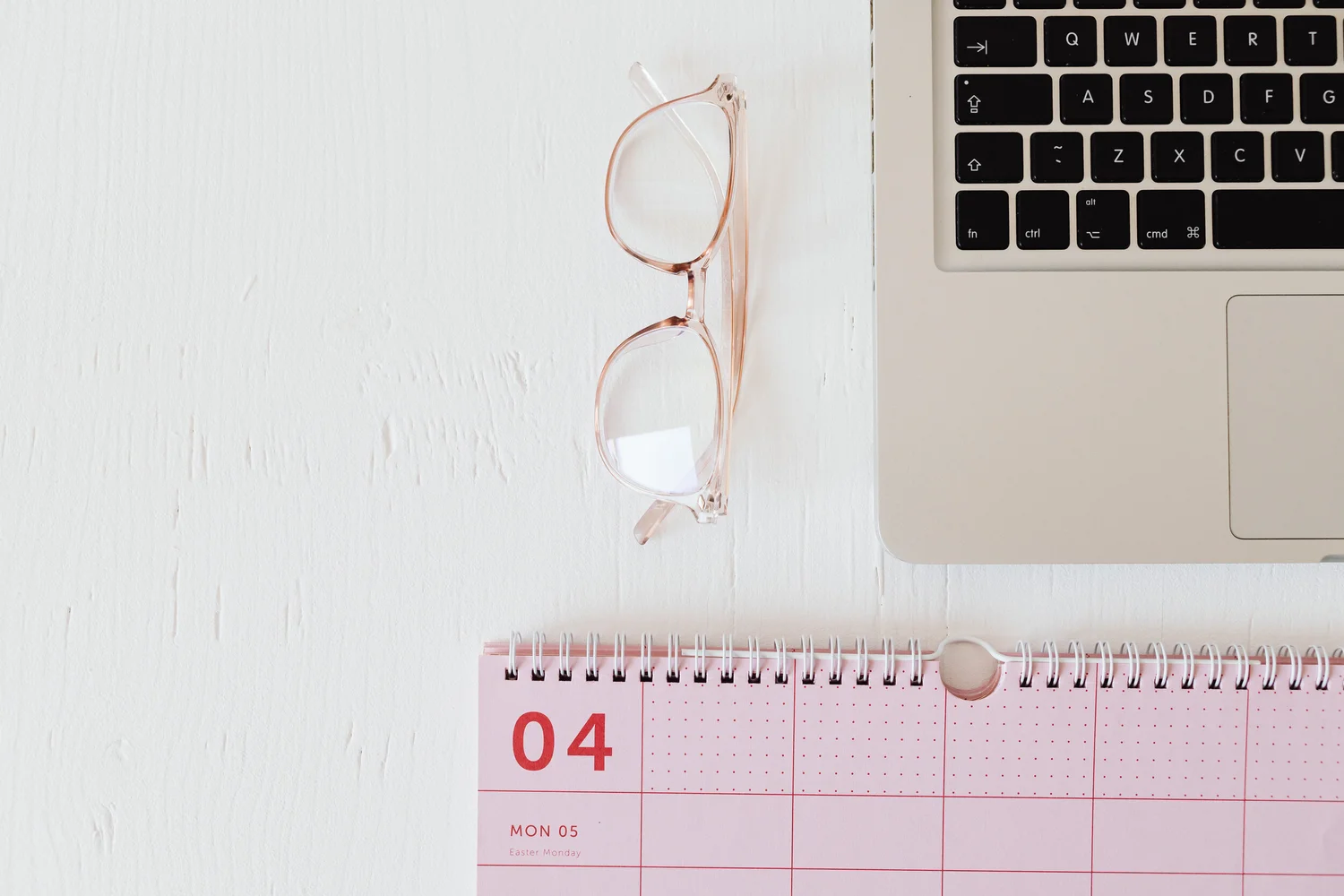 Glasses, a pink desk calendar and a laptop on a white desk