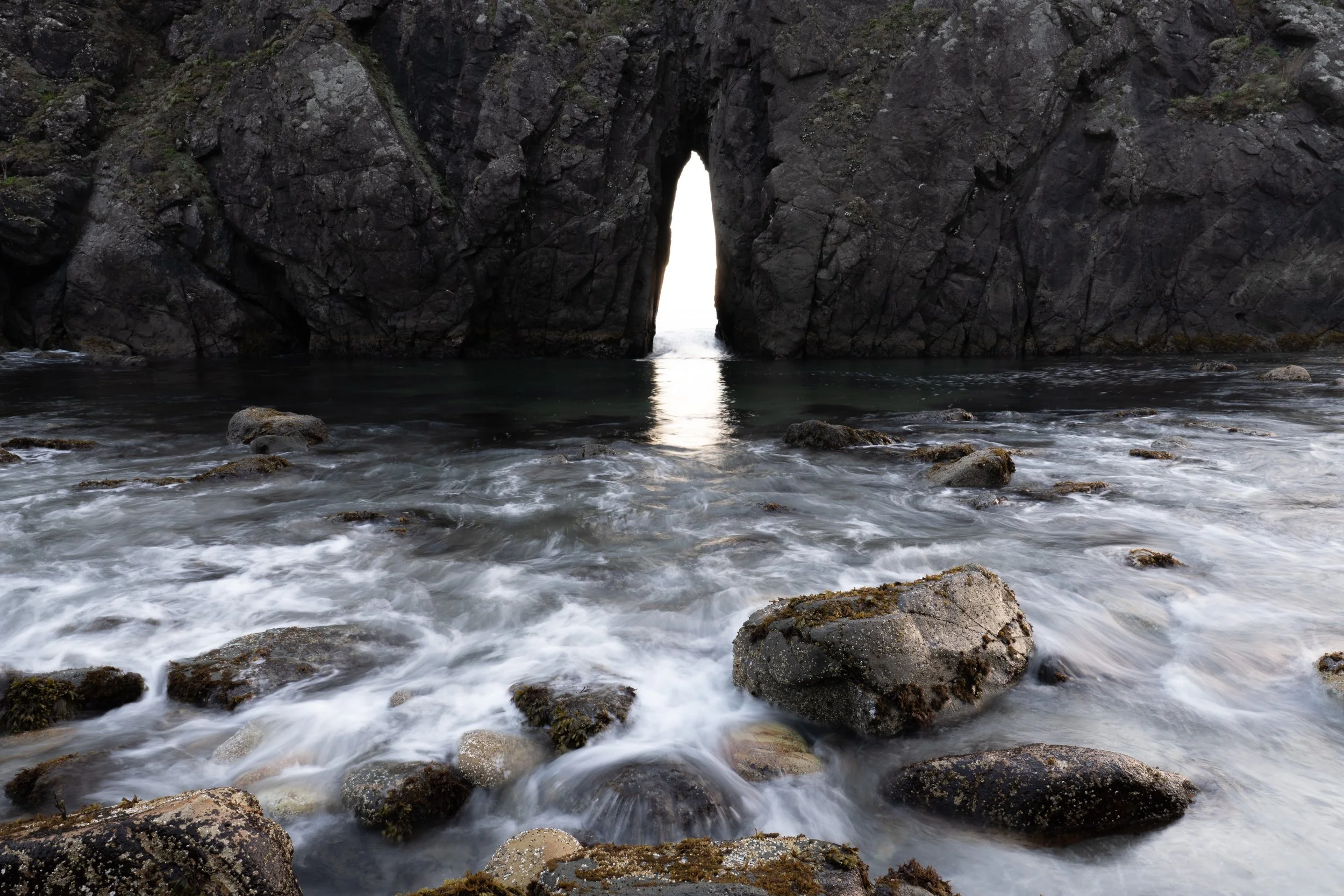 Ocean waves crashing over rocks in foreground with large rock formation and sea cave opening in background.
