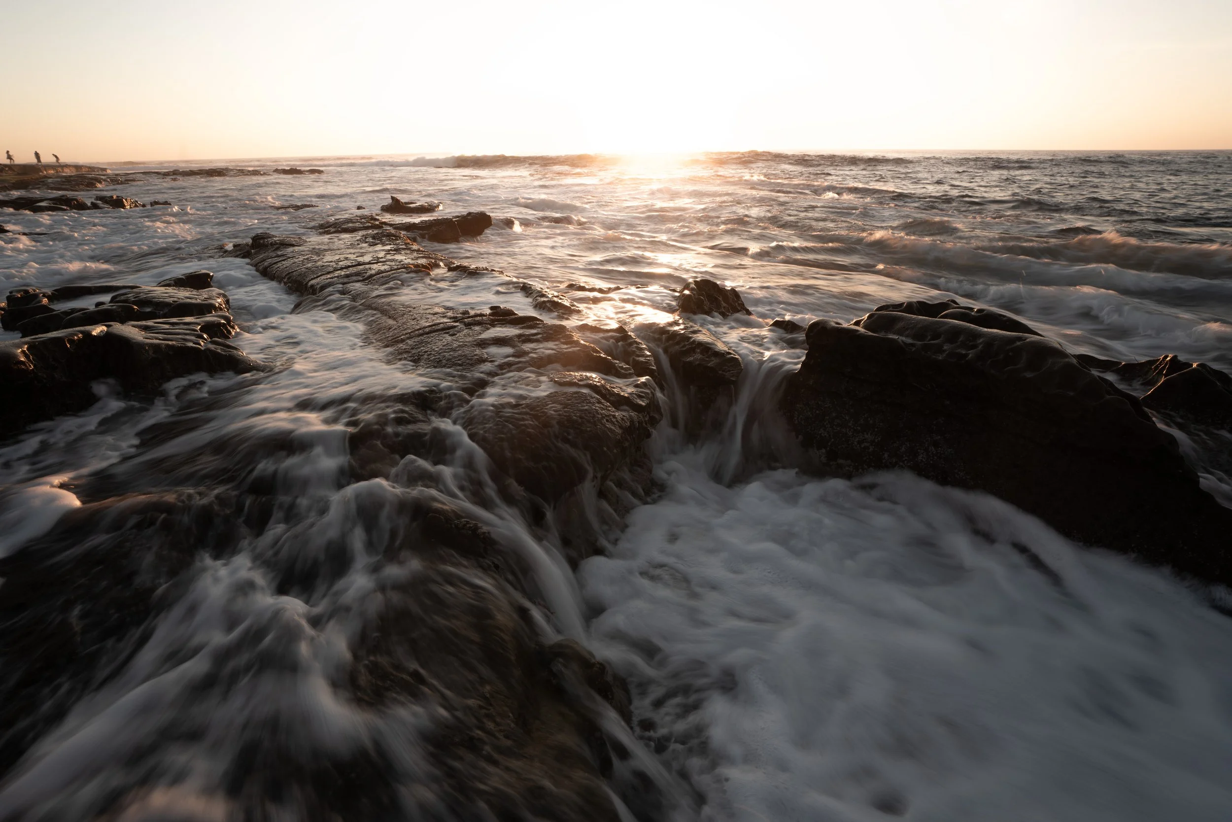 Sunset over rocky ocean shore with waves crashing and foam, with a few people in the distance on the coast.