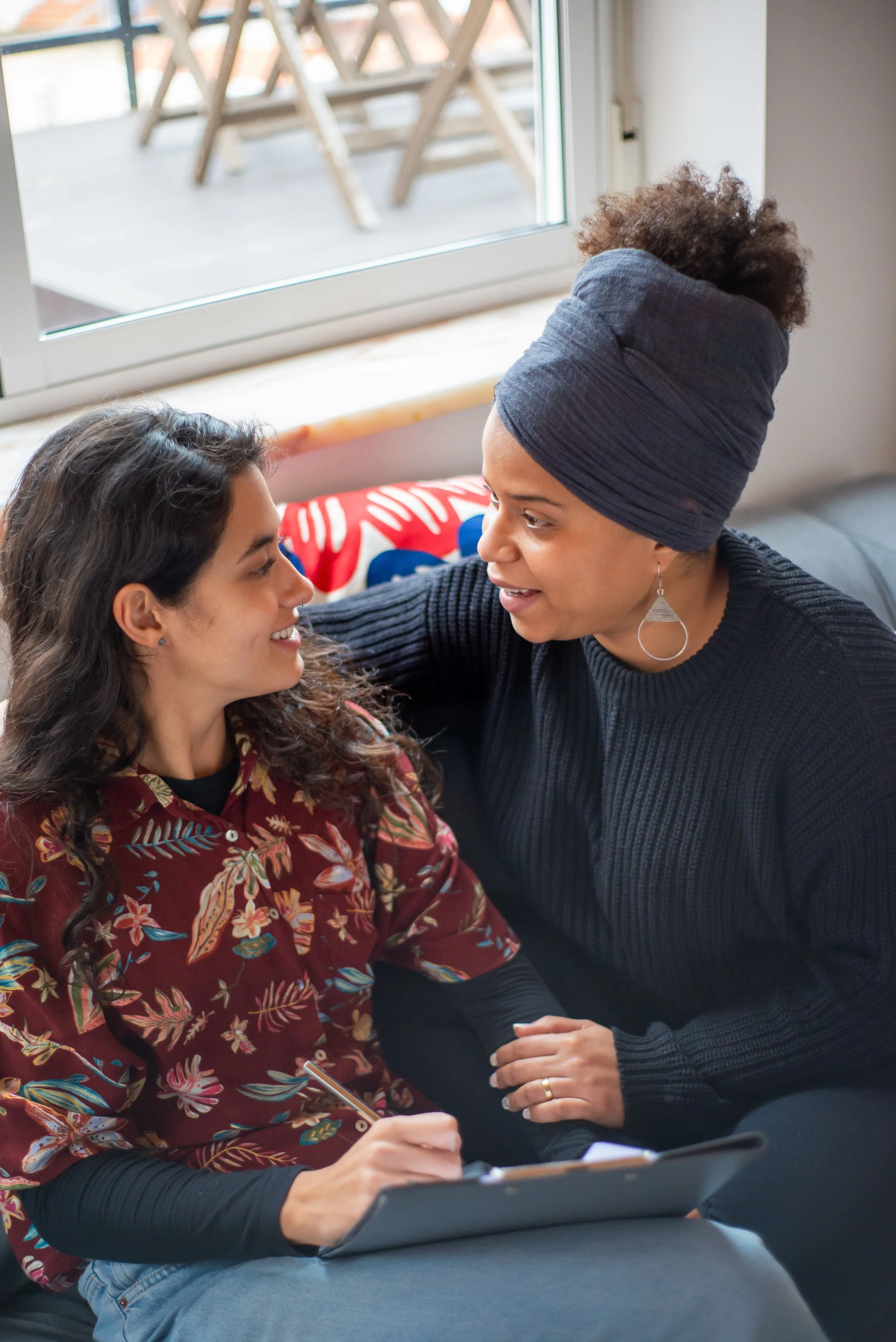 Two women sitting close together, engaging in a conversation. The woman on the left has long, curly dark hair and is holding a pencil and notebook. The woman on the right has short curly hair wrapped in a dark headwrap, is wearing earrings and a black sweater. They are indoors near a window, with a balcony visible outside.