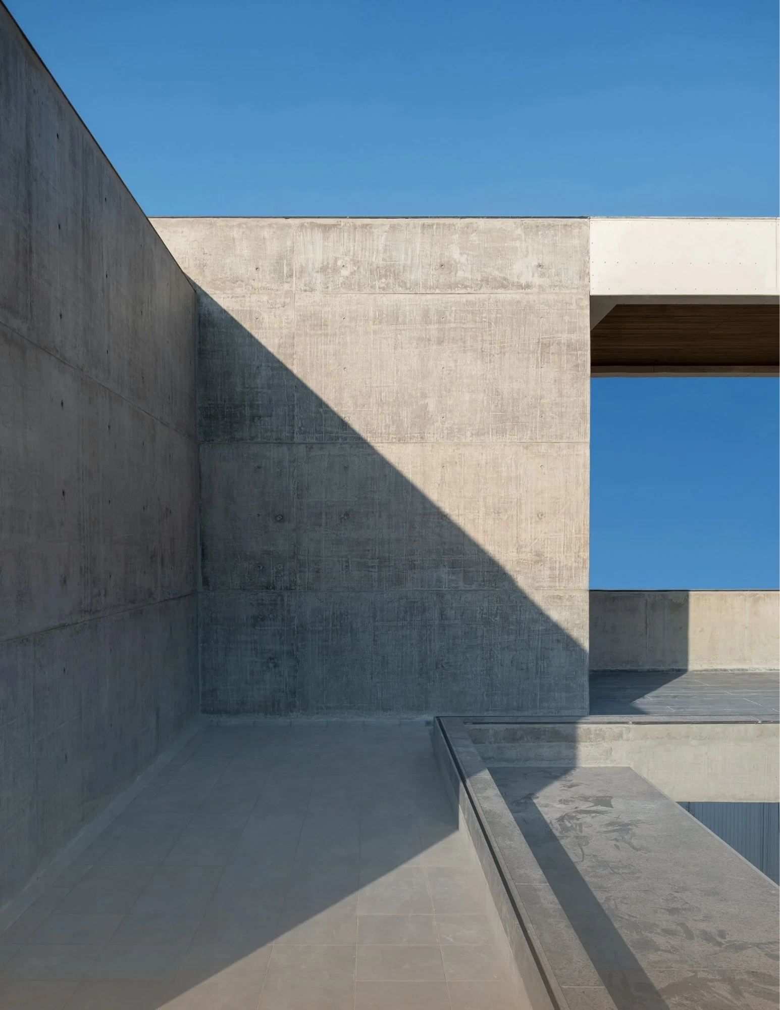 Modern concrete building with geometric shapes and sharp angles, shadow cast on the wall, clearing blue sky in the background.